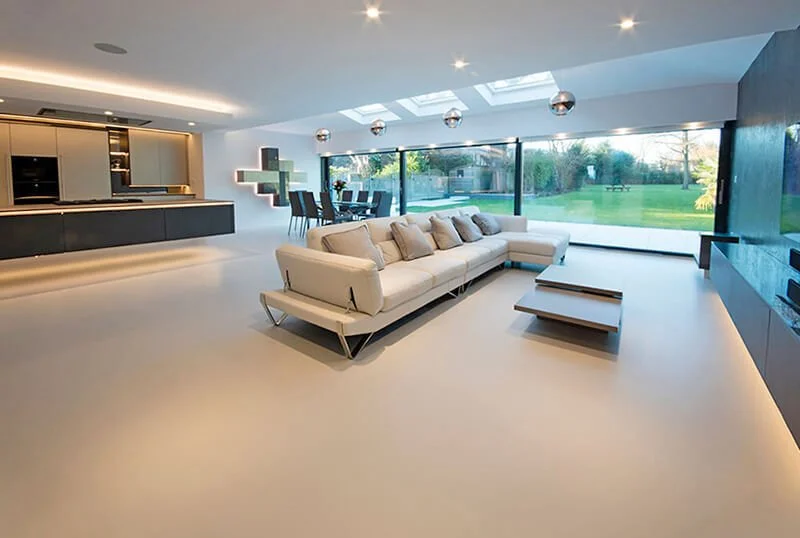 Modern open-concept living room with a large beige sectional sofa, low coffee tables, a black entertainment unit, and large windows overlooking a backyard with a pool and lawn.
