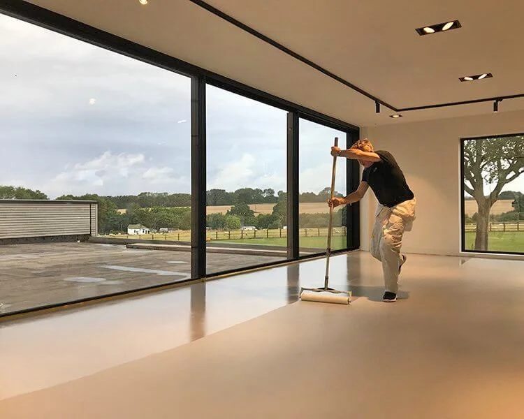 A worker skillfully applies a pigmented seal coat using a roller, transforming the large indoor floor into a stunning expanse of resin flooring, Hampshire UK.
