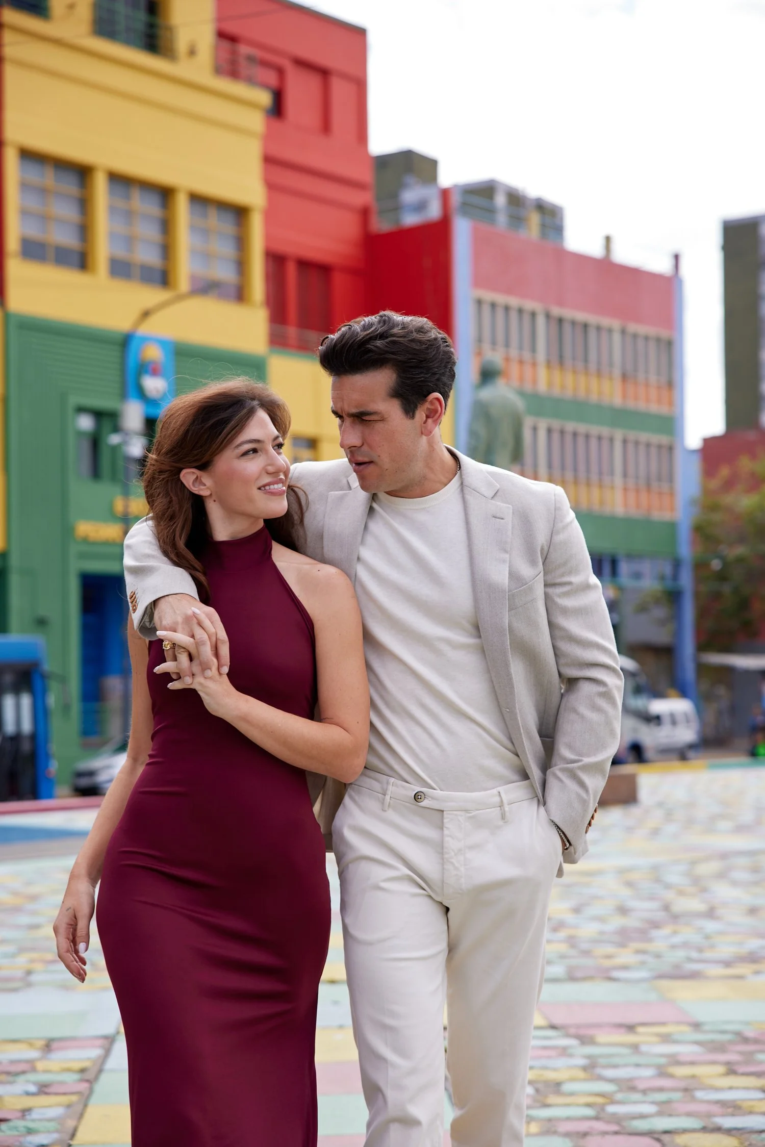 Una pareja caminando juntos en una calle colorida, con edificios en tonos amarillos, rojos y verdes al fondo. Ella lleva un vestido largo y morado, él viste un traje claro y camisa blanca.