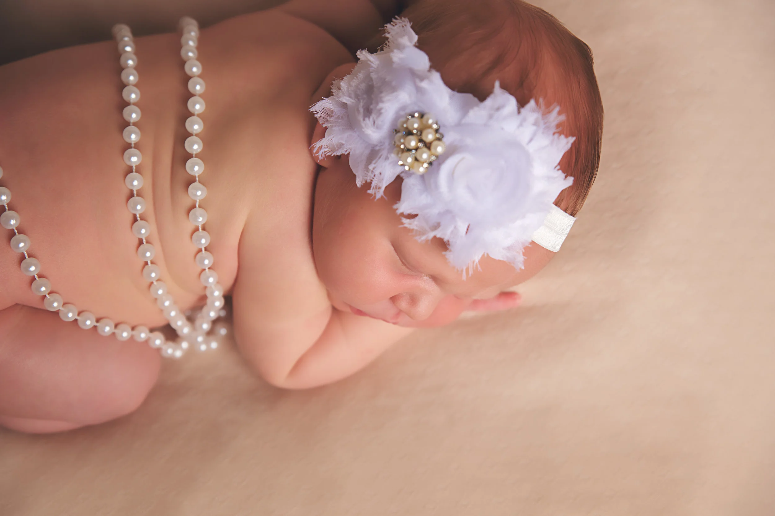 Newborn baby girl wearing a white floral headband and pearls during a professional newborn photography session.