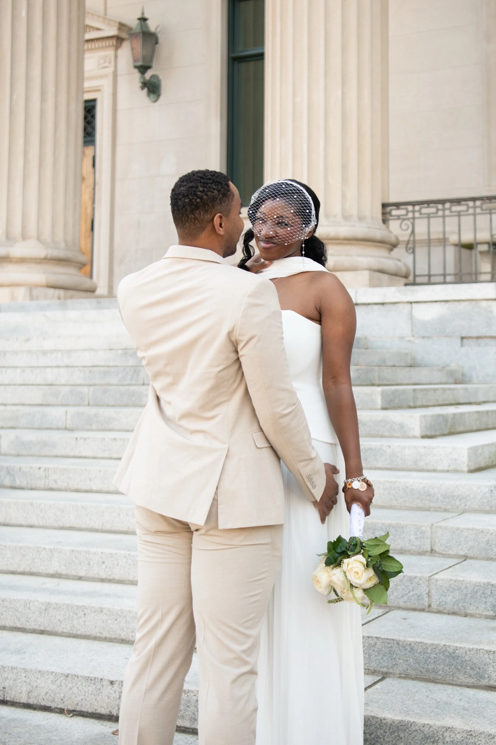 A couple dressed in wedding attire standing on steps in front of a historic building, with the woman holding a bouquet of flowers and wearing a birdcage veil.
