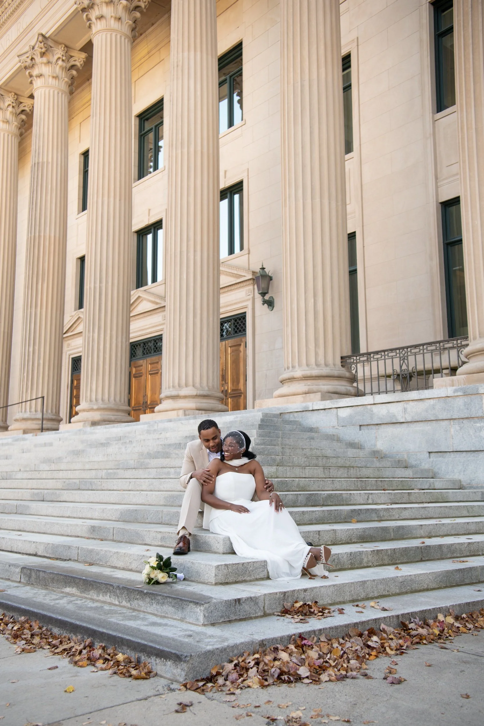 A newly married couple sitting on steps outside a grand neoclassical building with tall columns. The bride is wearing a strapless white wedding dress and veil, and the groom is in a light beige suit. A bouquet of white roses is placed on the steps ne