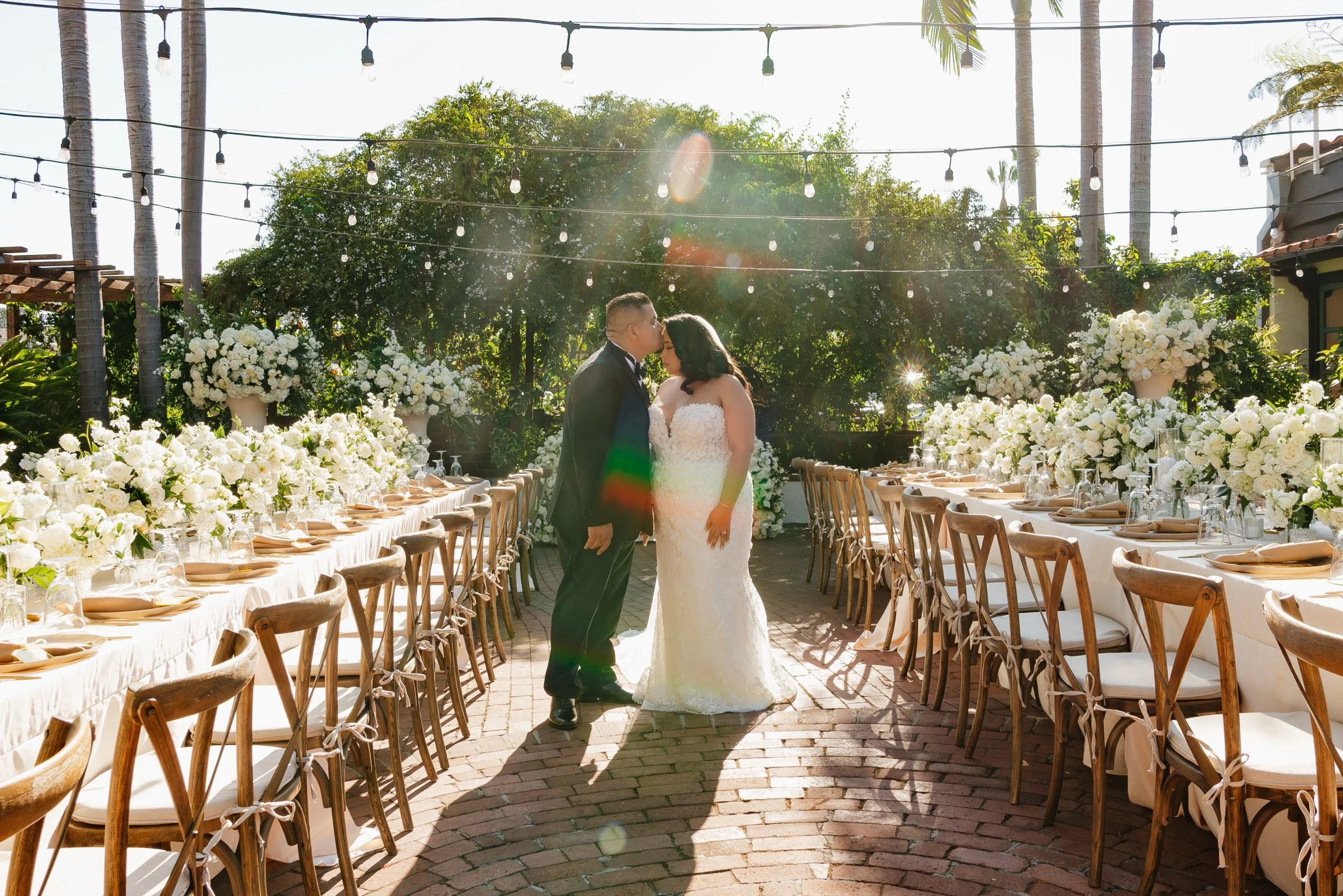 A newlywed couple sharing a kiss at a decorated outdoor wedding reception. The bride is in a white gown and the groom in a black tuxedo. Floral arrangements and dinner settings are on long tables on either side, with string lights overhead and lush g