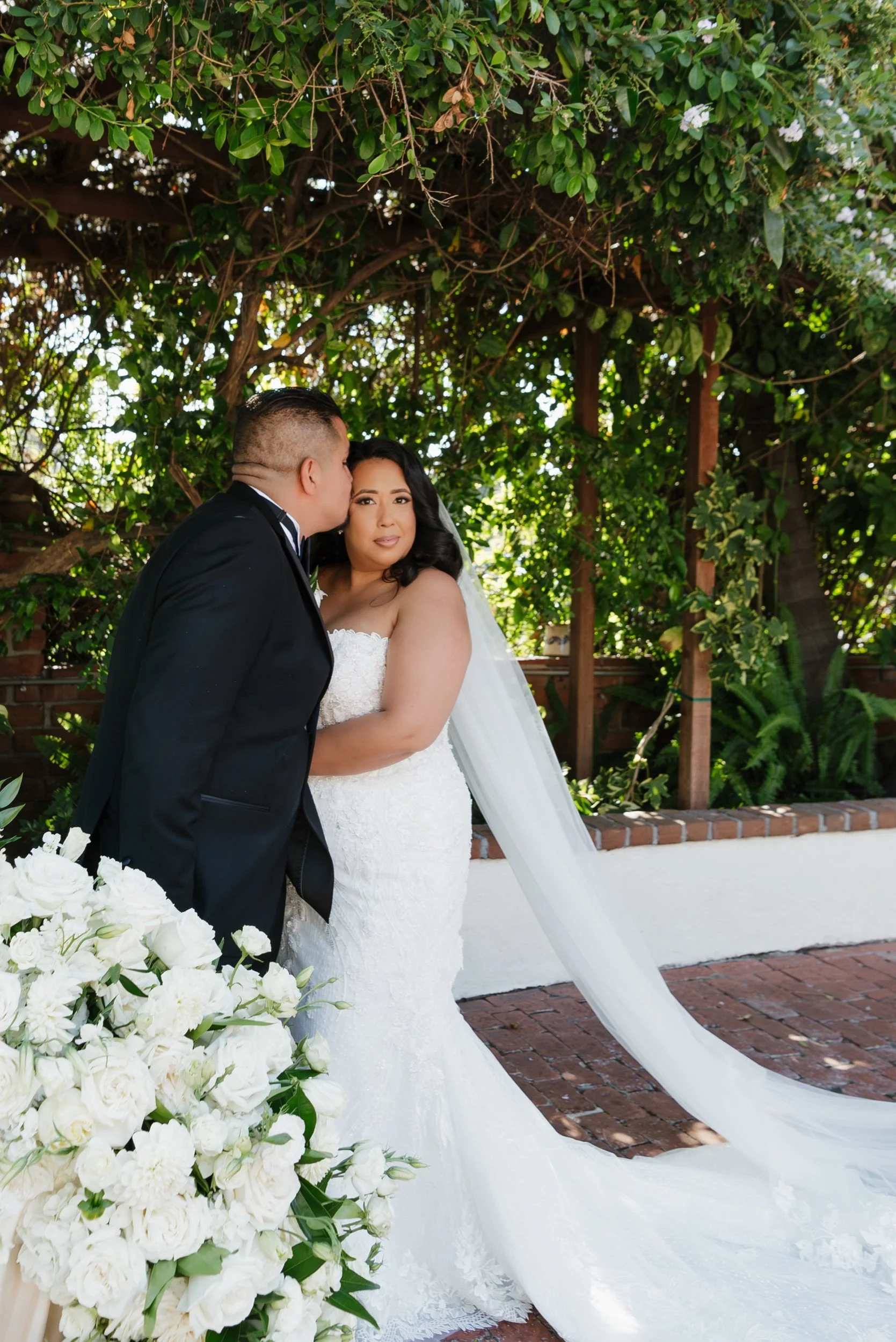 A newlywed couple in wedding attire, the groom kissing the bride on the cheek outdoors under lush greenery.