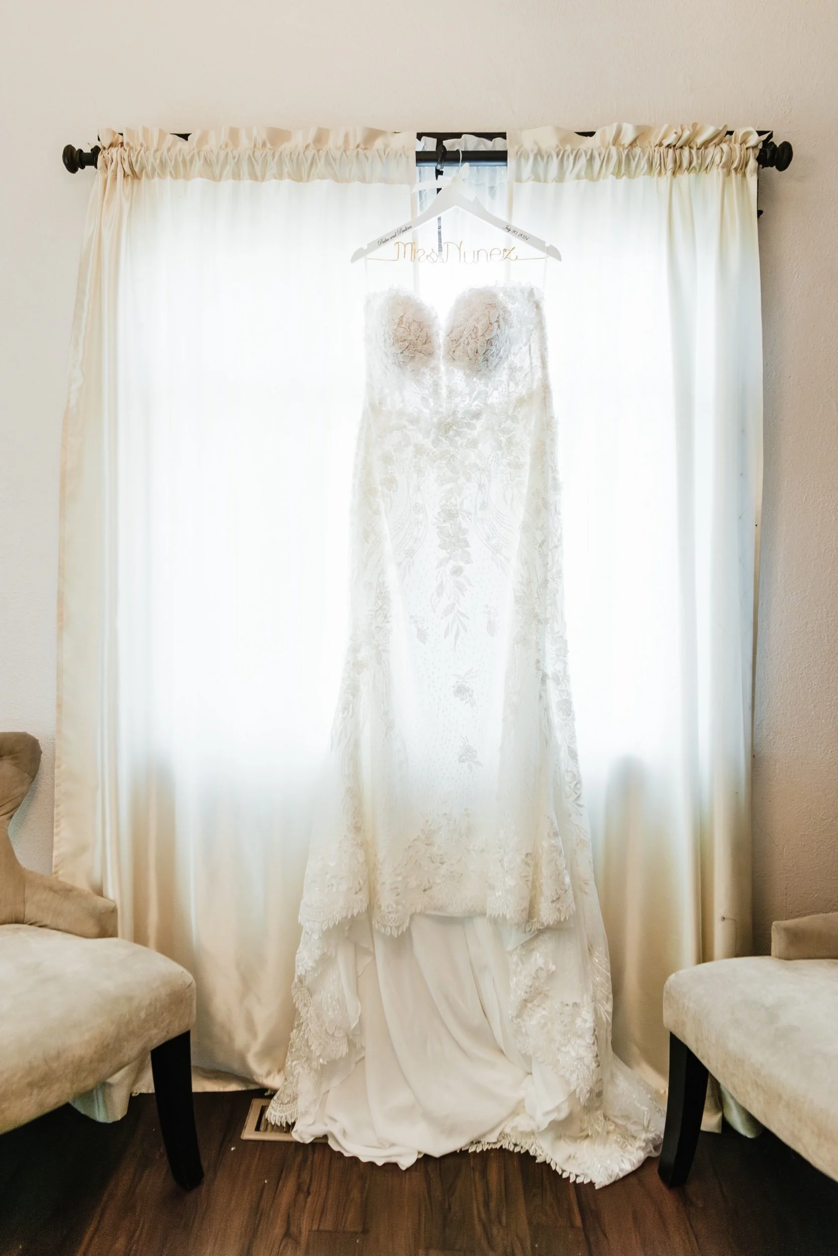 Wedding dress hanging on a hanger in front of a window with white curtains, bright backlighting creating a silhouette effect.