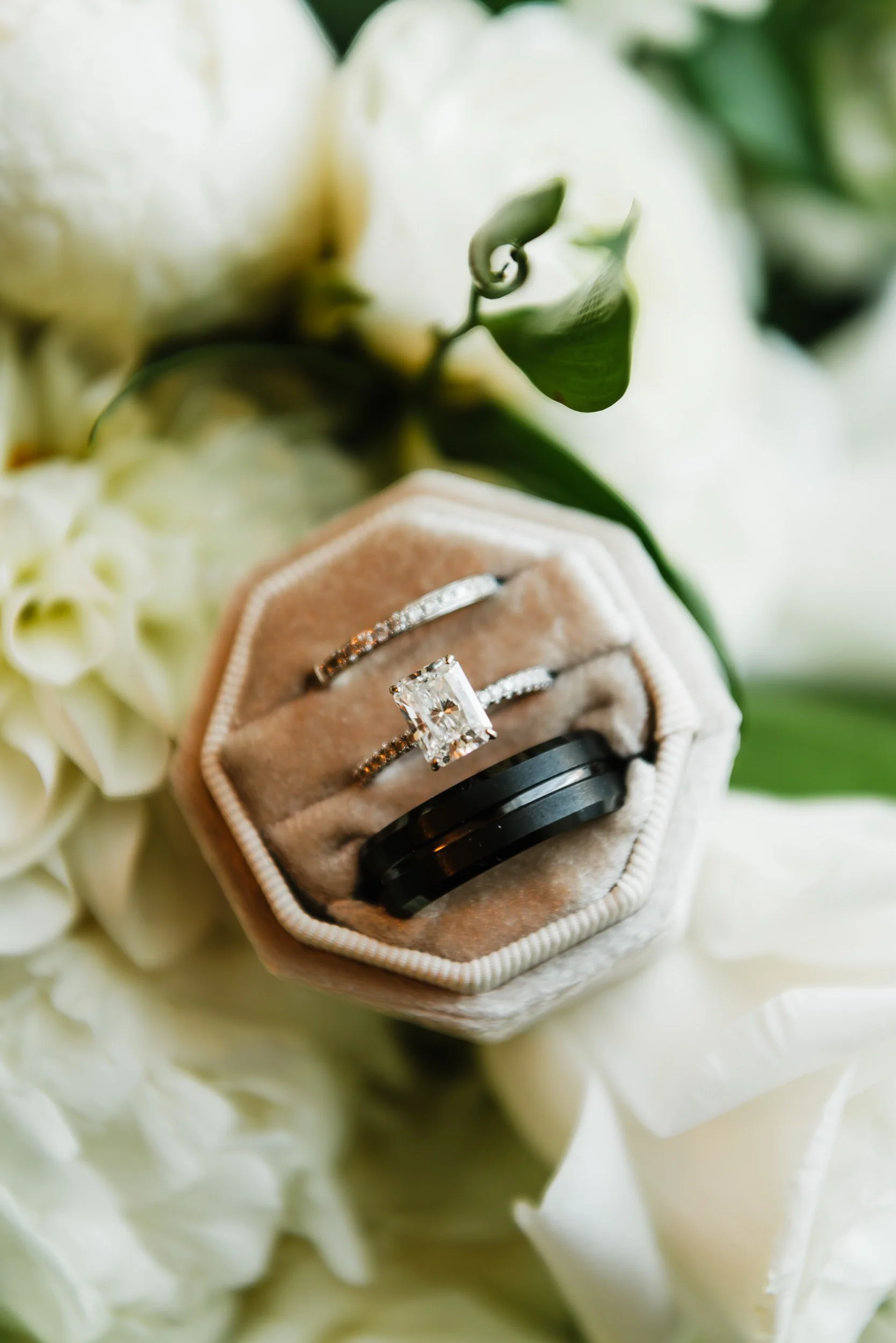 A close-up of a wedding ring box containing a diamond engagement ring with a princess-cut stone, two wedding bands, one with diamonds and the other plain black ceramic, surrounded by white flowers and green leaves.