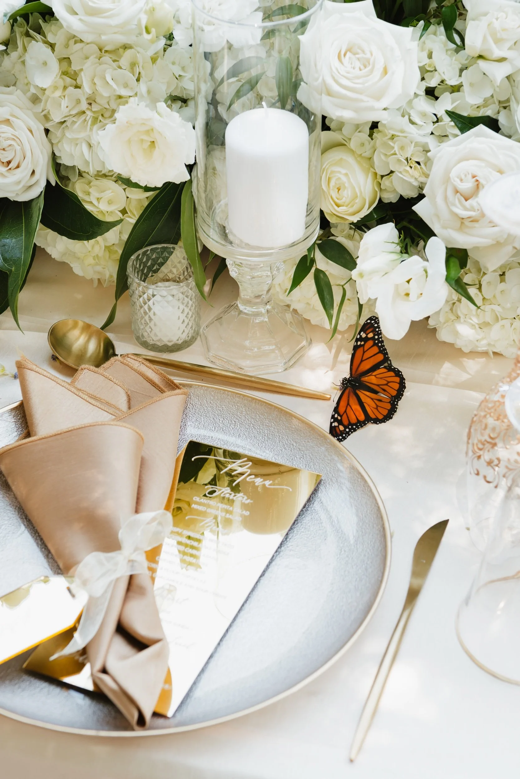 A wedding table setting with a large floral arrangement of white roses and hydrangeas. There is a white candle in a glass holder, a gold spoon, a gold napkin holder with a beige napkin tied with a white ribbon, and a gold-colored menu card with white