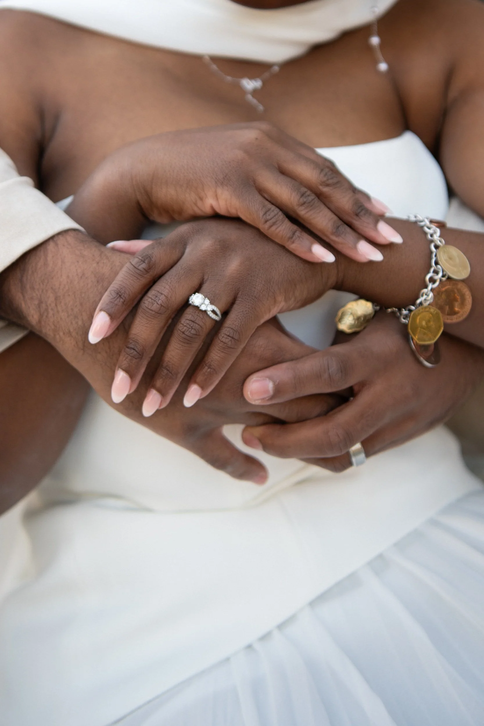 Close-up of a couple's hands, with the woman's hand resting on the man's arm. The woman wears a diamond ring, a bracelet with gold coins, and a necklace, while the man wears a wedding band. The background includes parts of their white clothing.