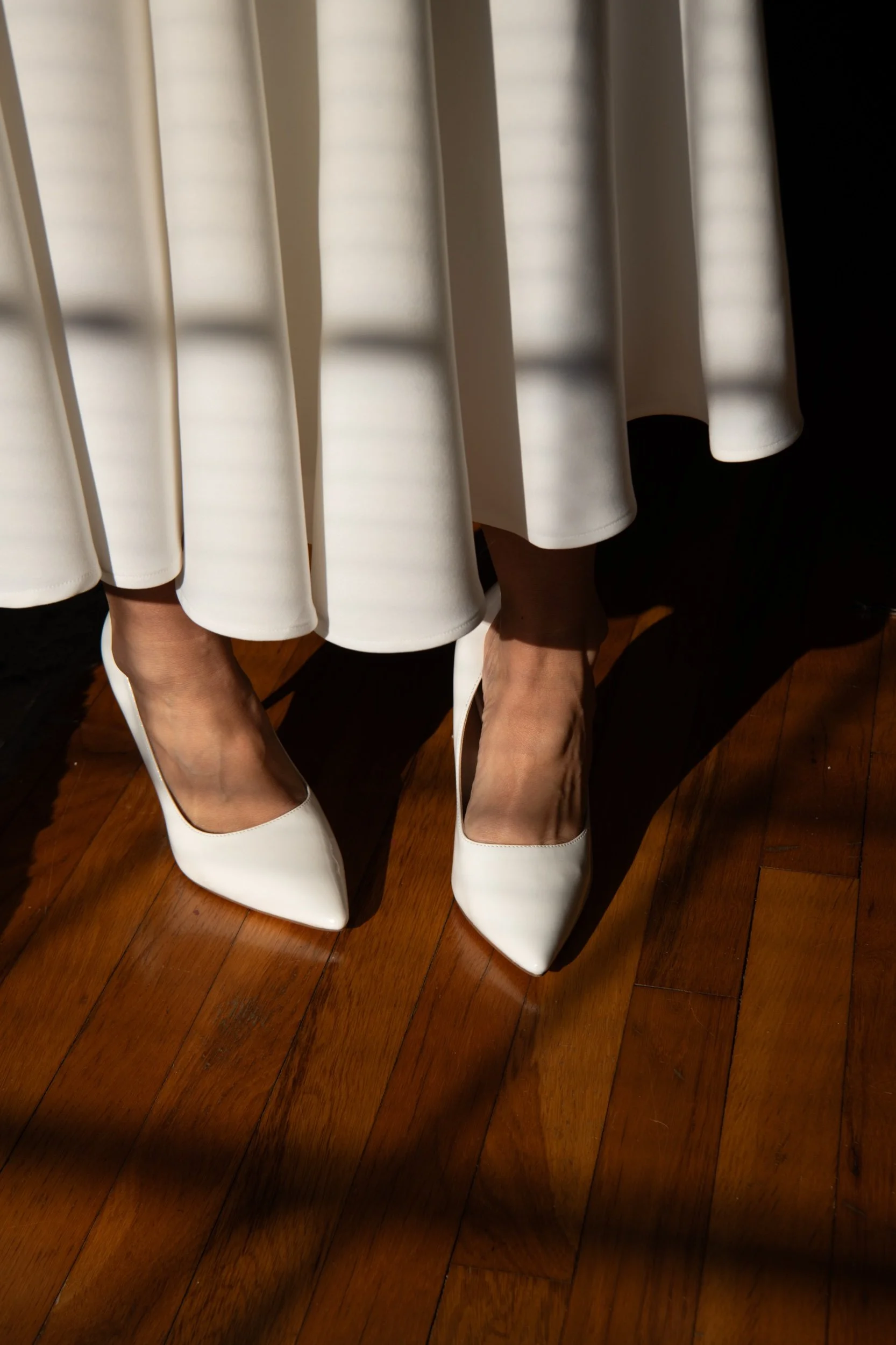 Close-up of a woman's legs and feet in white high-heeled shoes, wearing a white pleated skirt, standing on a wooden floor with sunlight casting shadows from window blinds.