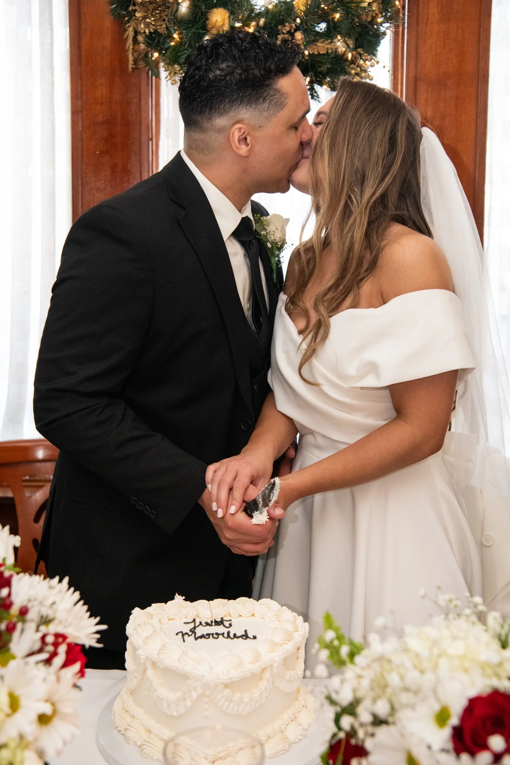 A bride and groom sharing a kiss during their wedding ceremony, holding hands, with a decorated wedding cake on the table and flowers in the foreground.