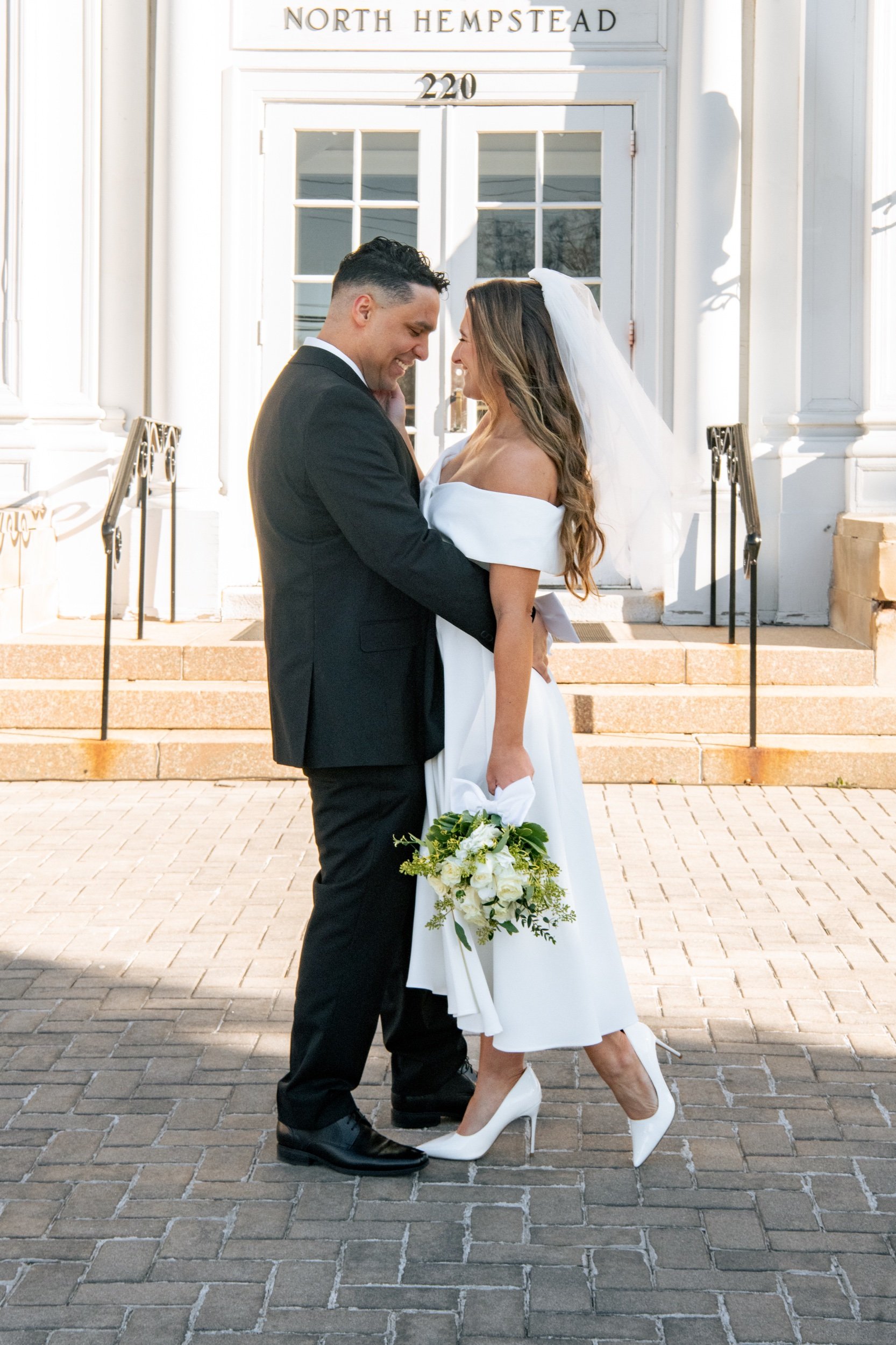 A bride and groom embracing in front of a white building with steps. The bride holds a bouquet of white flowers, and herself wears a white dress with off-the-shoulder sleeves and white high heels. The groom is dressed in a black suit and black shoes.