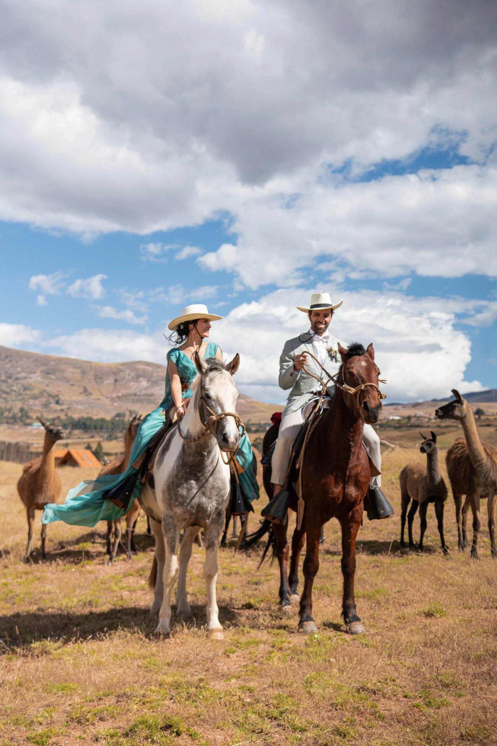 A man and woman riding horses in a rustic outdoor setting with mountains and a cloudy sky, surrounded by llamas.