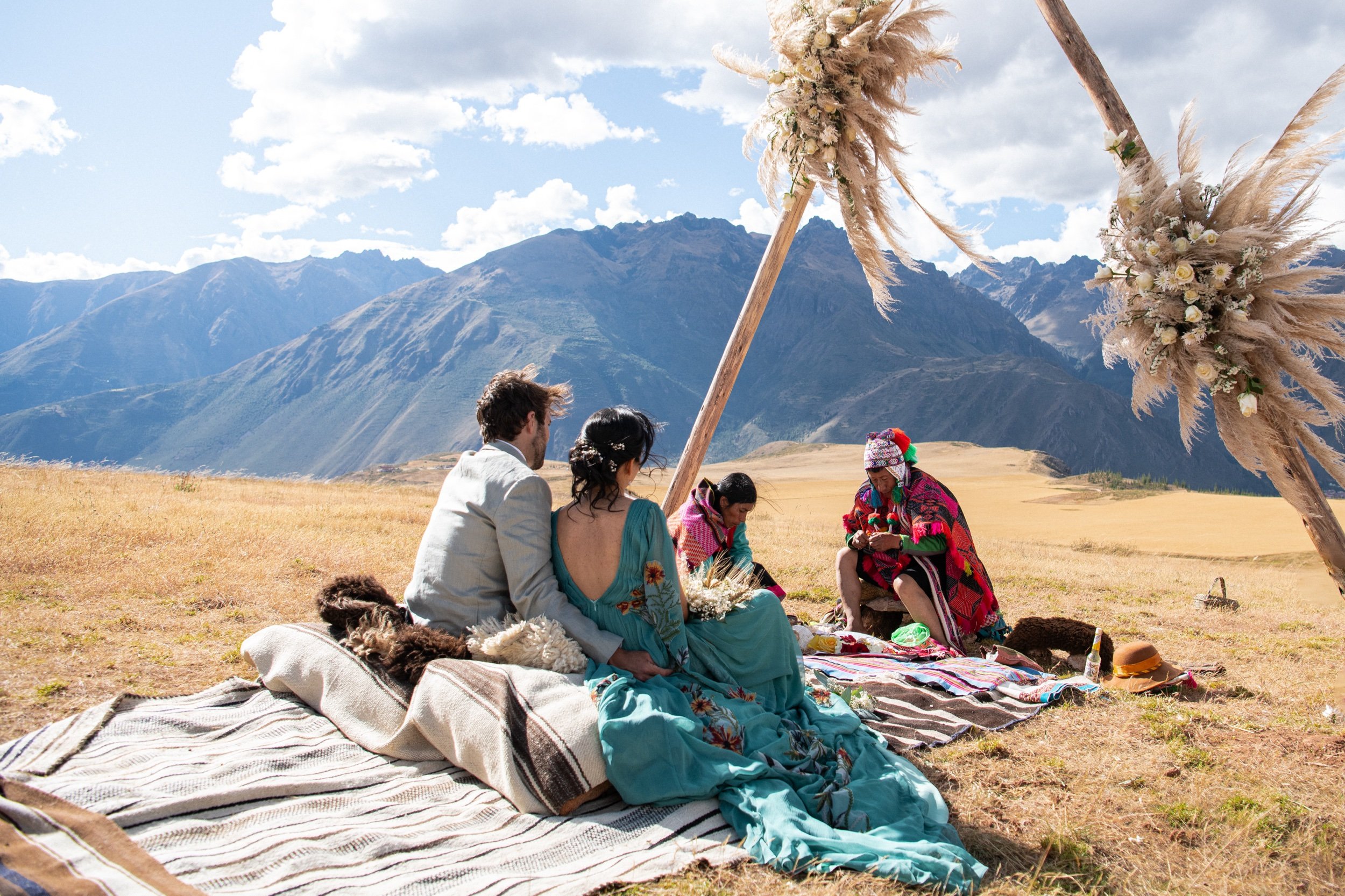 A couple dressed in wedding attire sitting on a blanket in a grassy field, with a group of indigenous women in traditional clothing performing a ceremony under large decorated wooden structures, with mountains in the background.