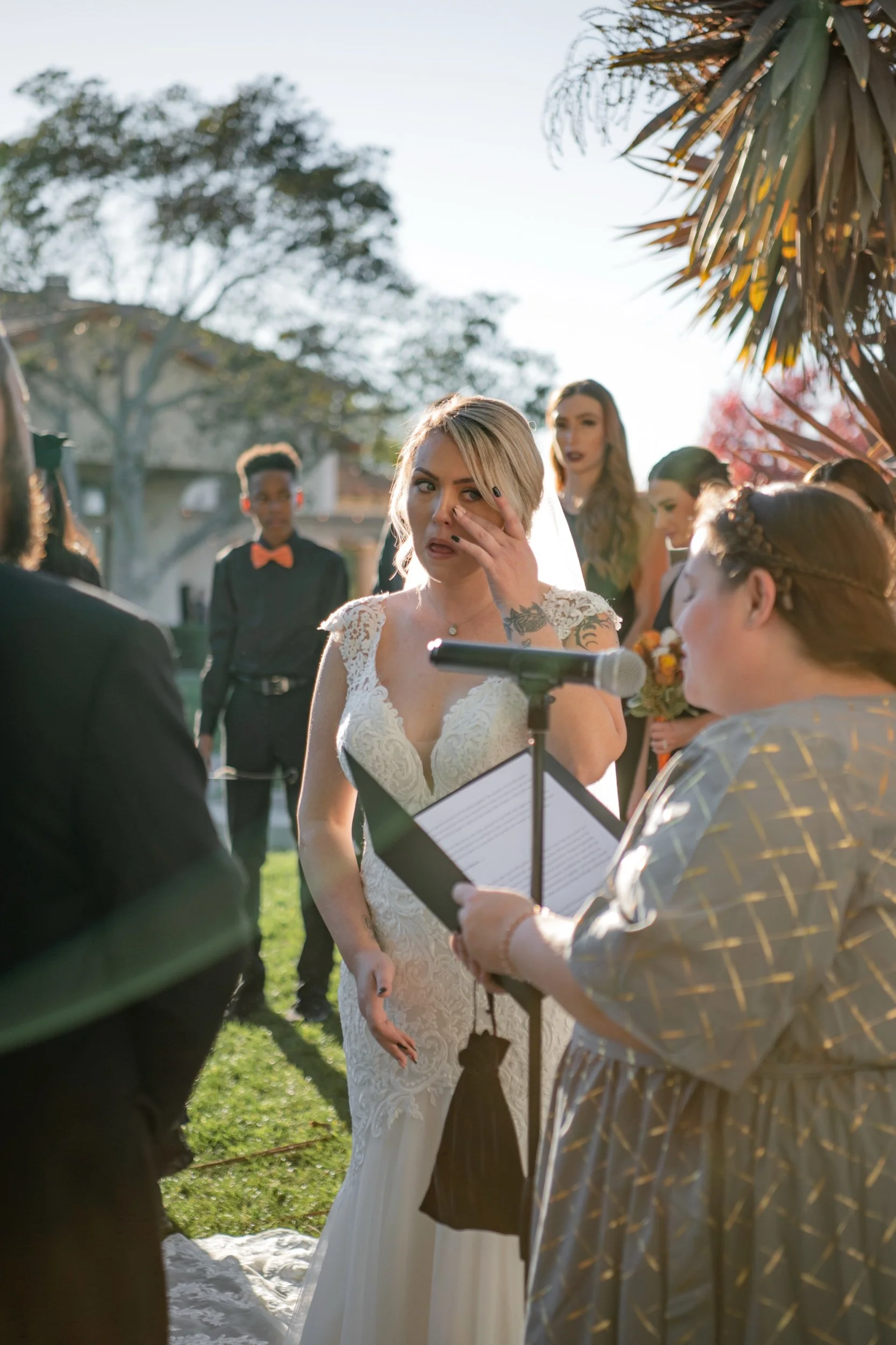 A bride with blonde hair, wearing a white lace wedding gown, wiping tears during her wedding ceremony outdoors, surrounded by bridesmaids and guests, with sunlight and trees in the background.