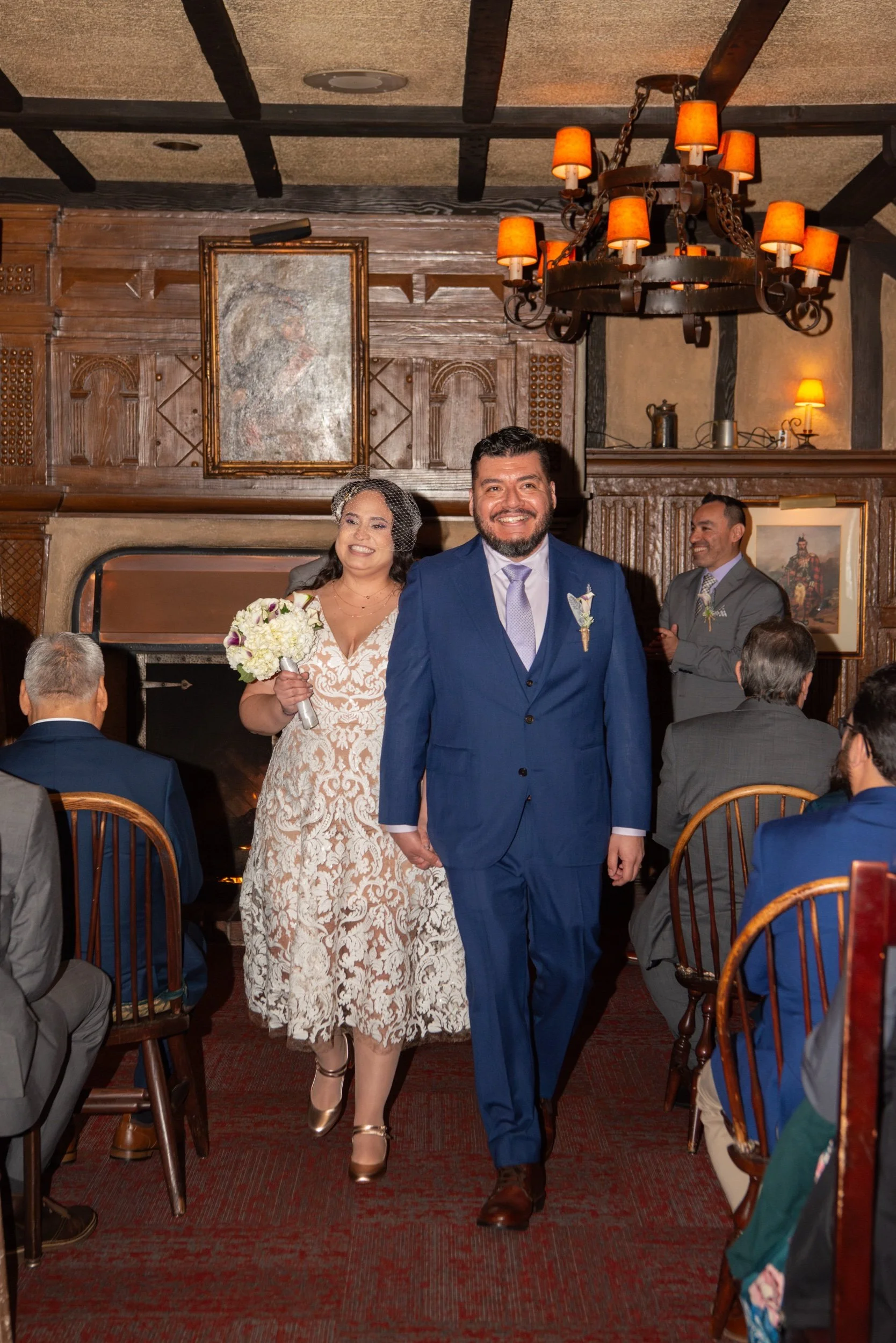 A bride and groom walking down the aisle at a wedding reception in a warmly lit room with wooden walls and a fireplace. The bride is holding a bouquet and wearing a lace dress, while the groom is dressed in a blue suit.