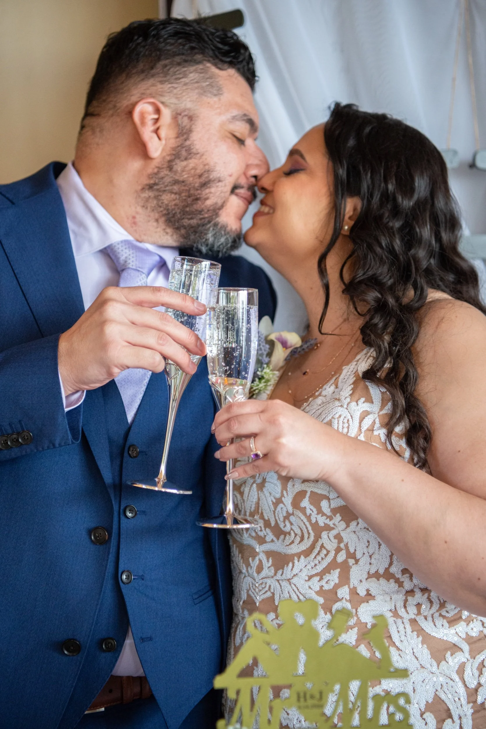 A newlywed couple, a man in a blue suit and a woman in a white lace dress, share a kiss while holding champagne flutes during their wedding celebration.