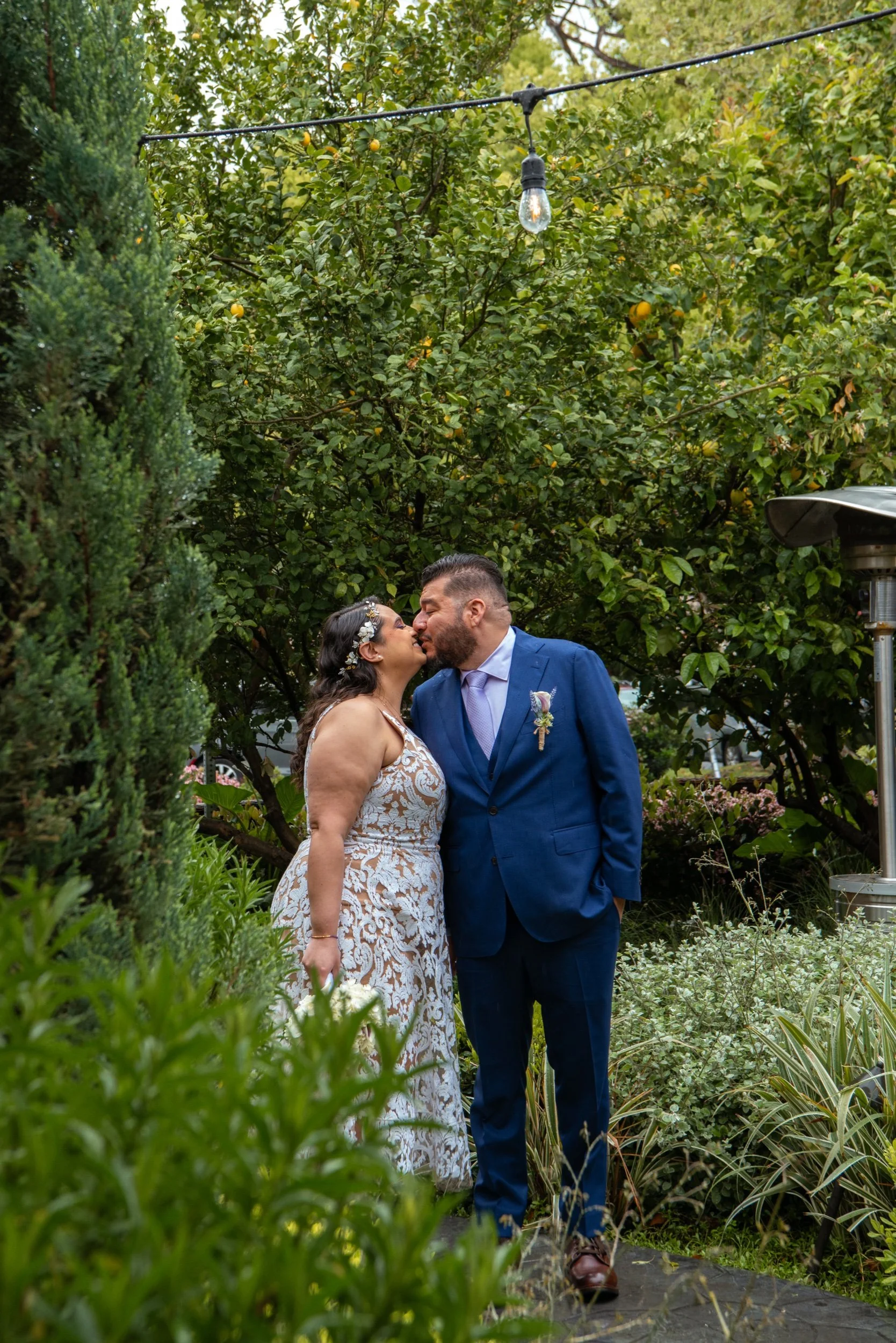 A couple dressed in wedding attire sharing a kiss in a lush garden with green trees and plants, hanging lights overhead, and a patio heater on the right.