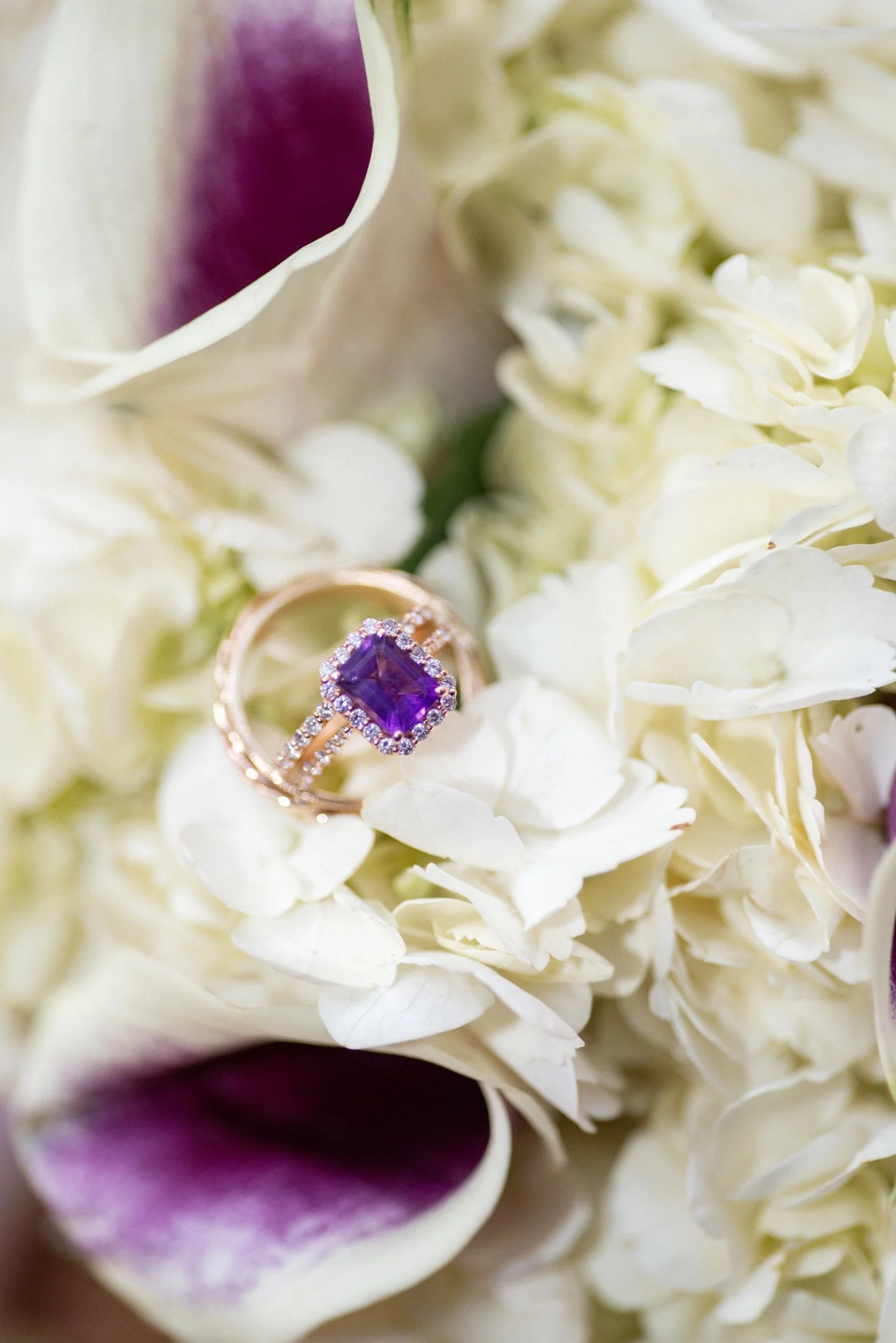 A close-up of an engagement ring with a large purple gemstone surrounded by small diamonds, placed on white flowers with purple accents.
