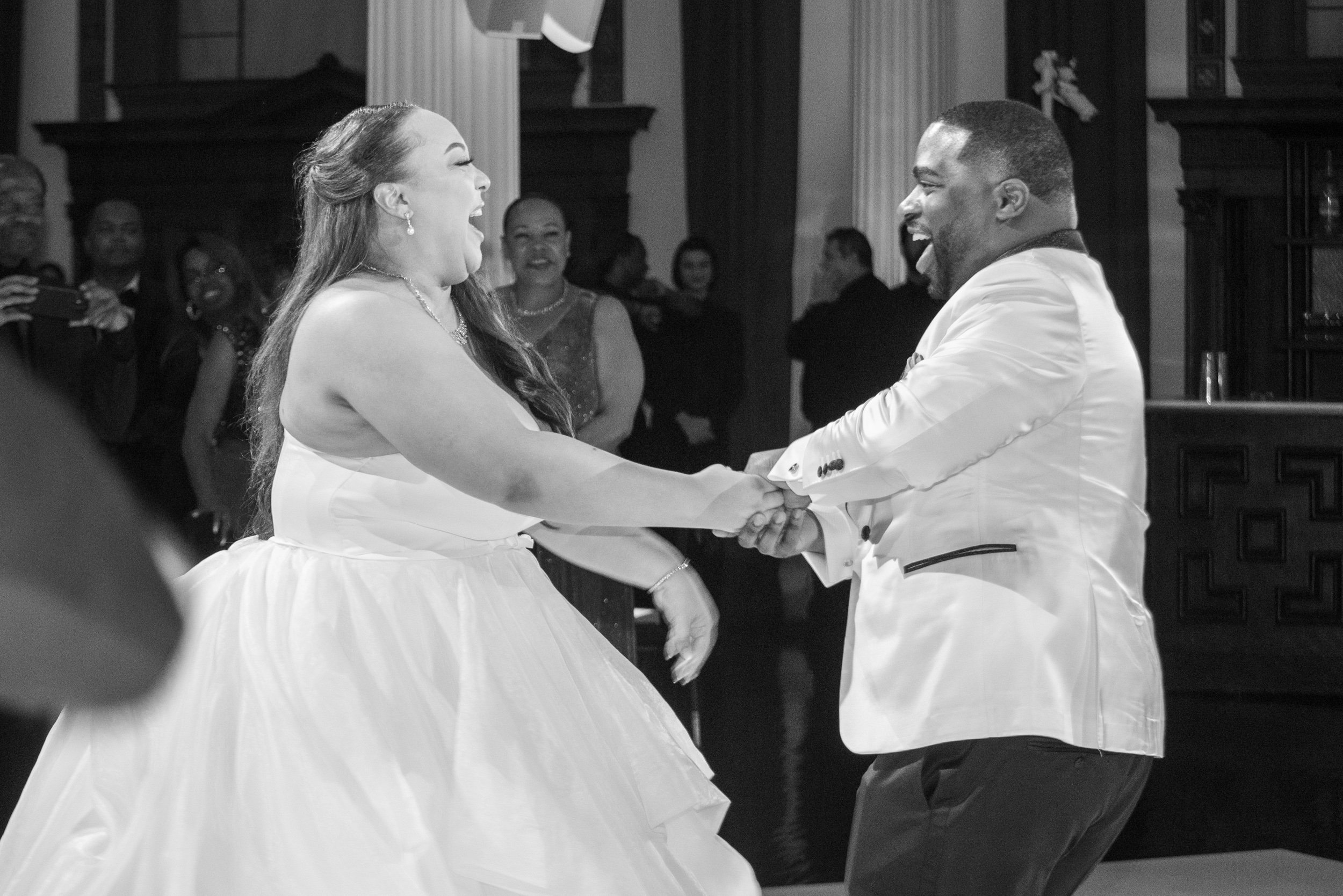A joyful couple dancing at a wedding reception, holding hands and laughing, with friends watching and smiling in the background.