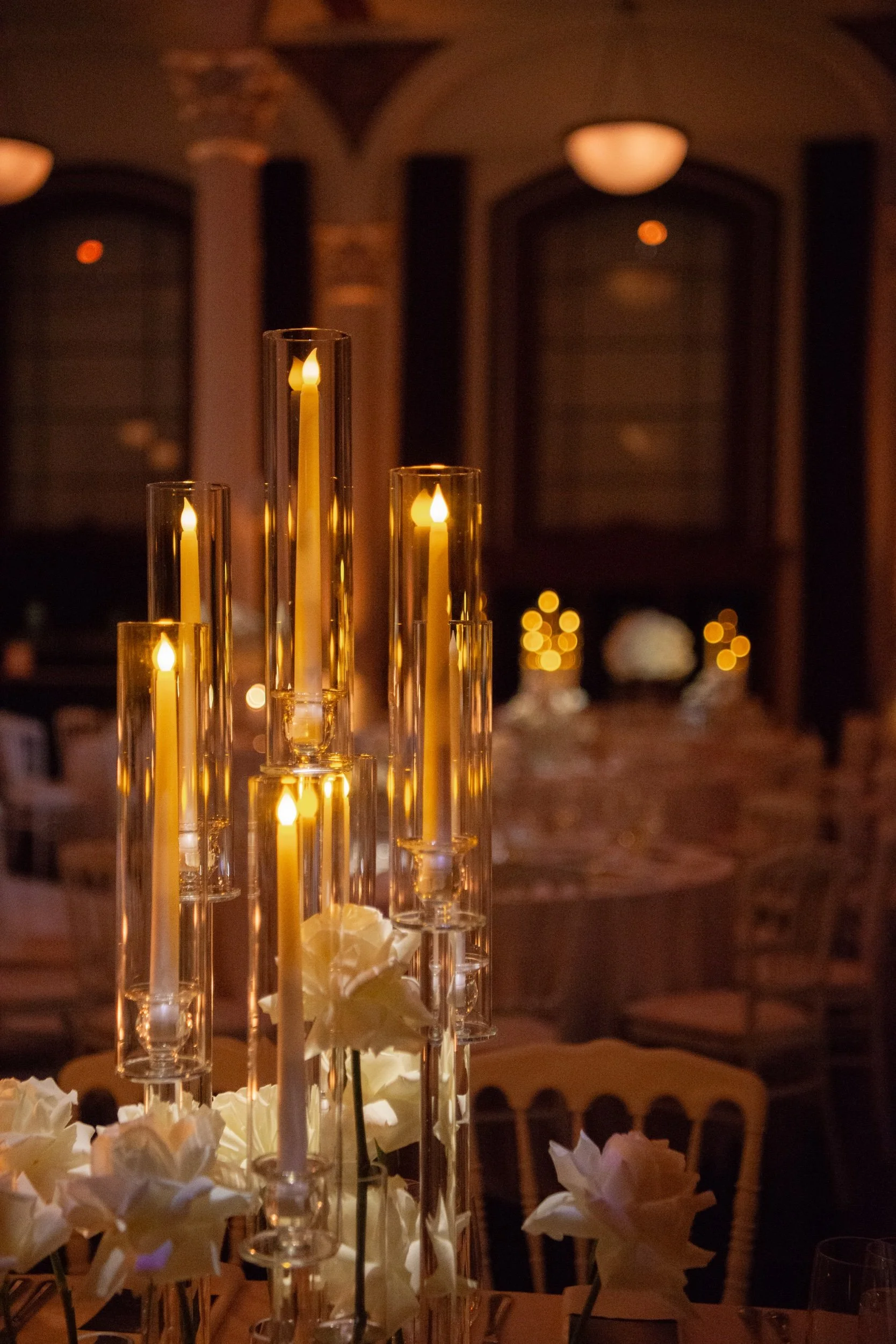 Elegant dining table decorated with tall candleholders with lit candles and white flowers, in a warmly lit room.
