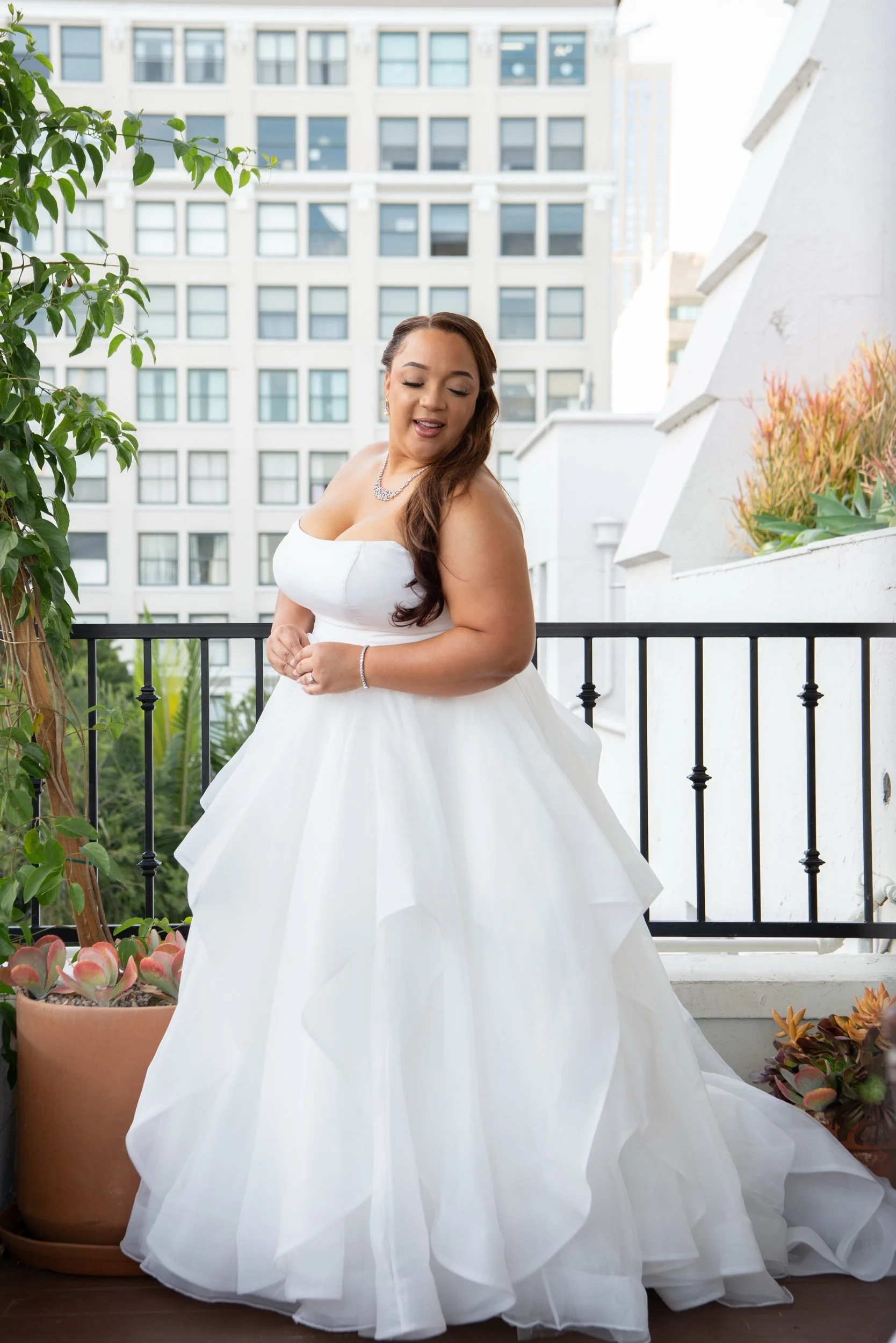 A bride in a white gown stands on a balcony with potted plants and metal railing, with city buildings in the background.