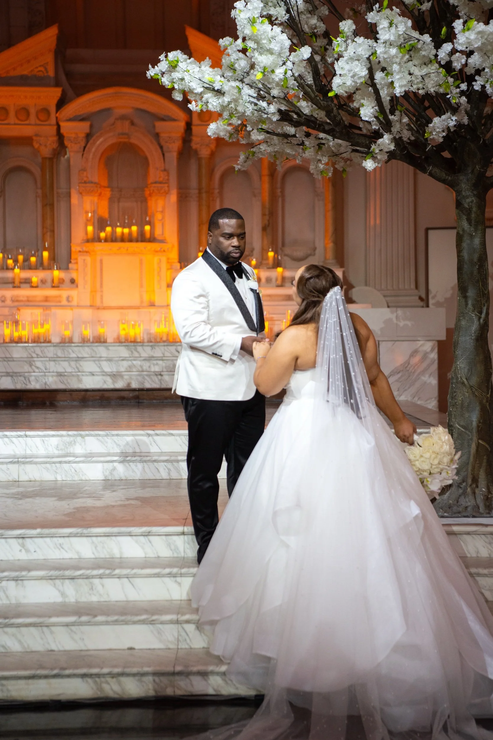A couple exchanging vows during a wedding ceremony inside a church, with the bride wearing a white wedding dress and veil, and the groom in a white tuxedo jacket with black lapels, next to a decorated tree with white flowers.