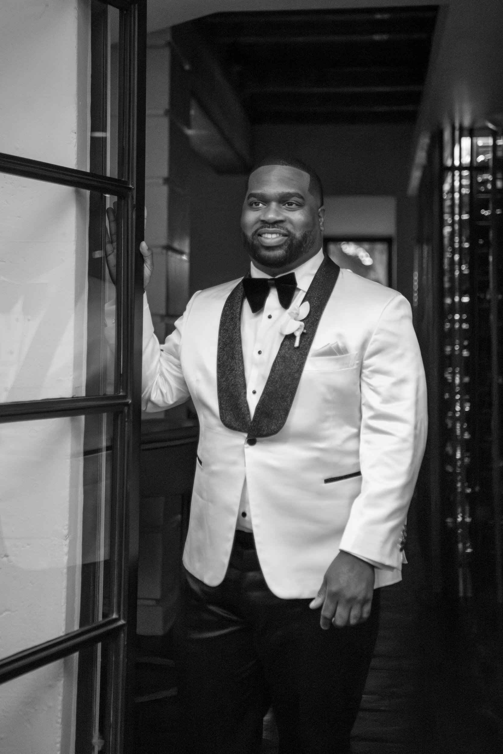 A man dressed in a tuxedo with a white jacket, black bow tie, and boutonniere, smiling and standing by a door.