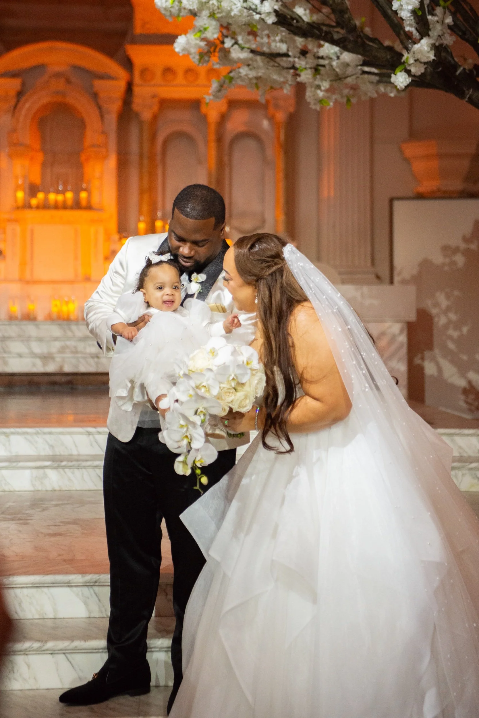 A bride and groom holding a baby during their wedding ceremony in a church, with the bride in a white gown and veil, the groom in a light-colored tuxedo, and an altar with candles in the background.