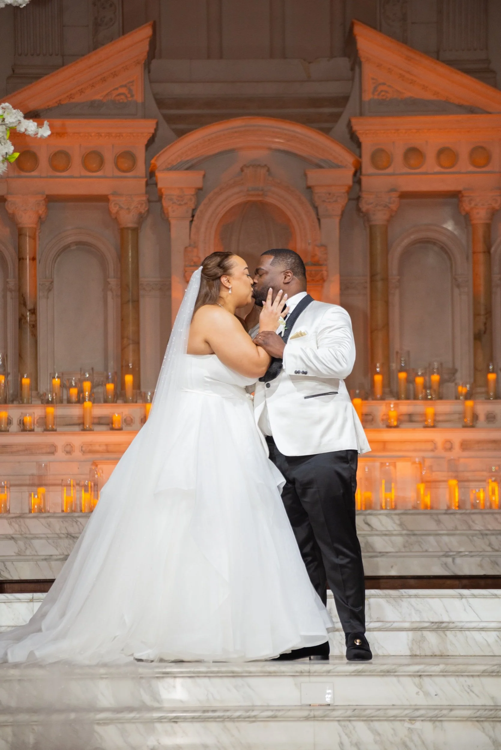 A bride and groom kissing during their wedding ceremony, standing on a marble staircase with a background of lit candles and an ornate architectural setting.
