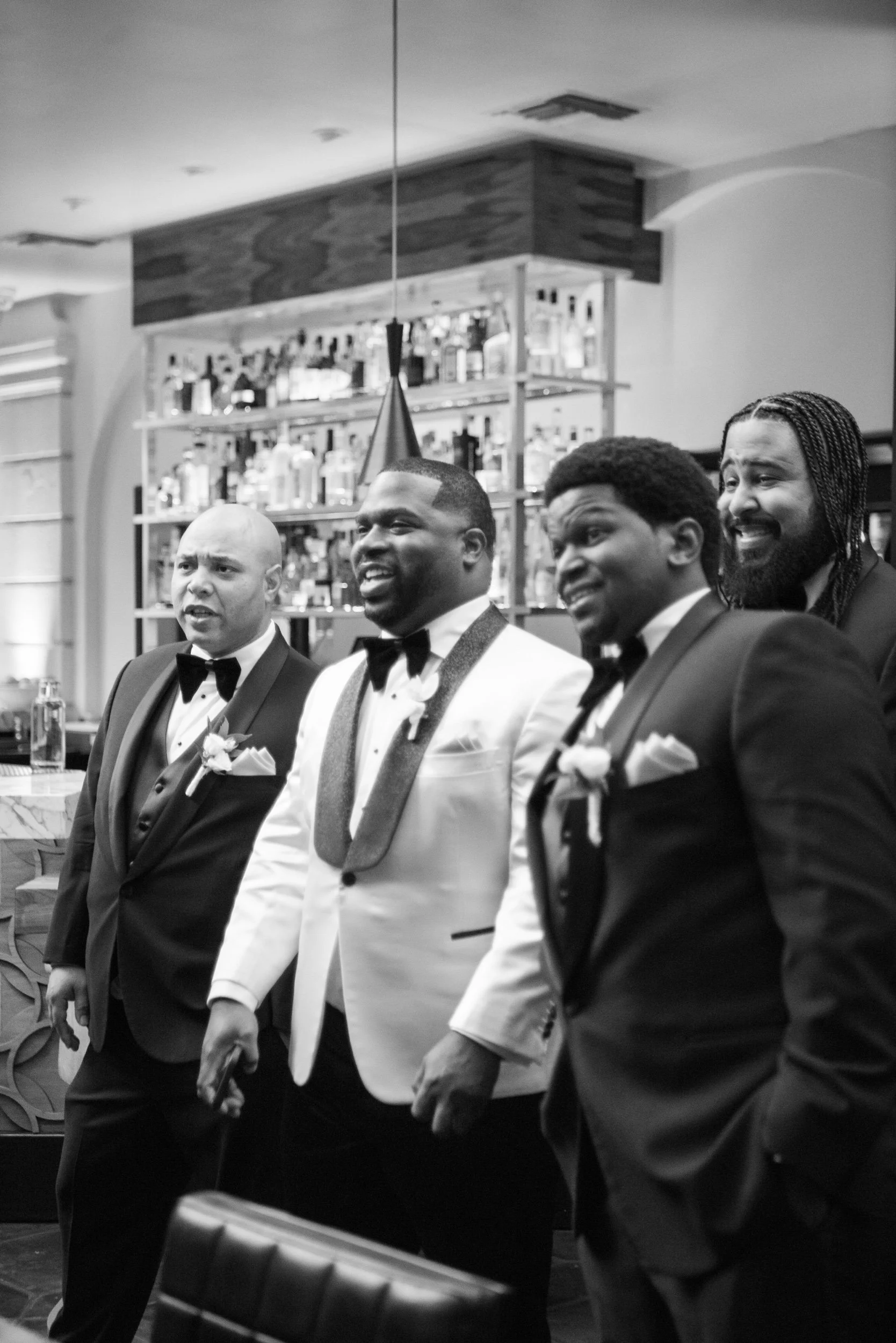 Four men in tuxedos standing in a bar or restaurant, some with boutonnières, with bottles on shelves behind them, appearing to be at a formal event or wedding celebration.