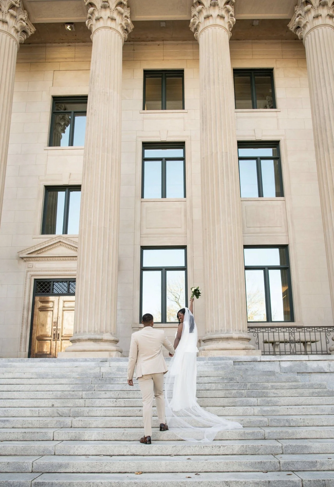 A bride and groom walking up stairs outside a neoclassical building with large columns, the bride holding a bouquet of flowers and raising her arm.