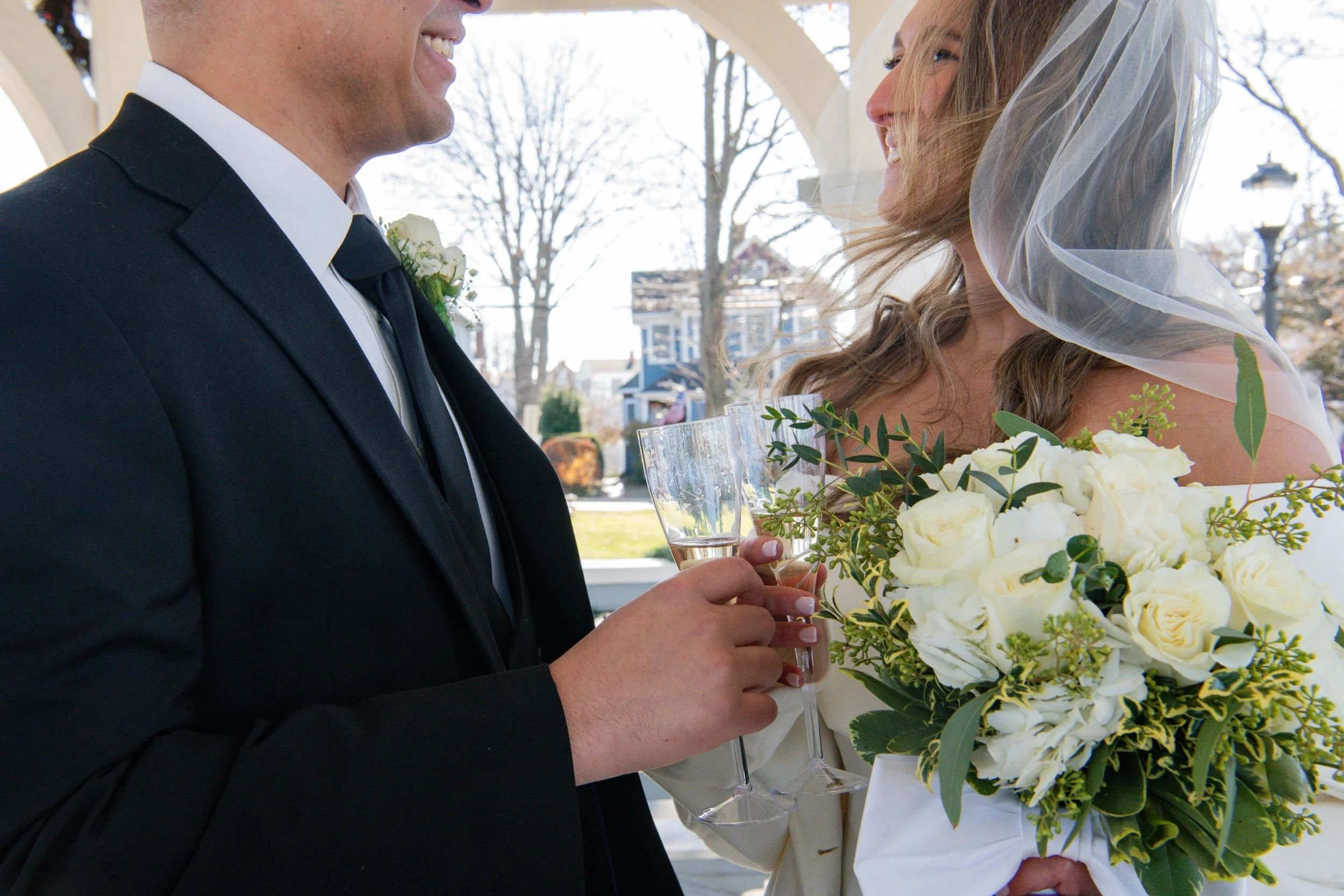 A wedding couple is smiling and raising glasses for a toast, with the bride holding a bouquet of white roses and greenery, and the groom wearing a black tuxedo with a white shirt and black tie. The bride has a veil and the background features outdoor