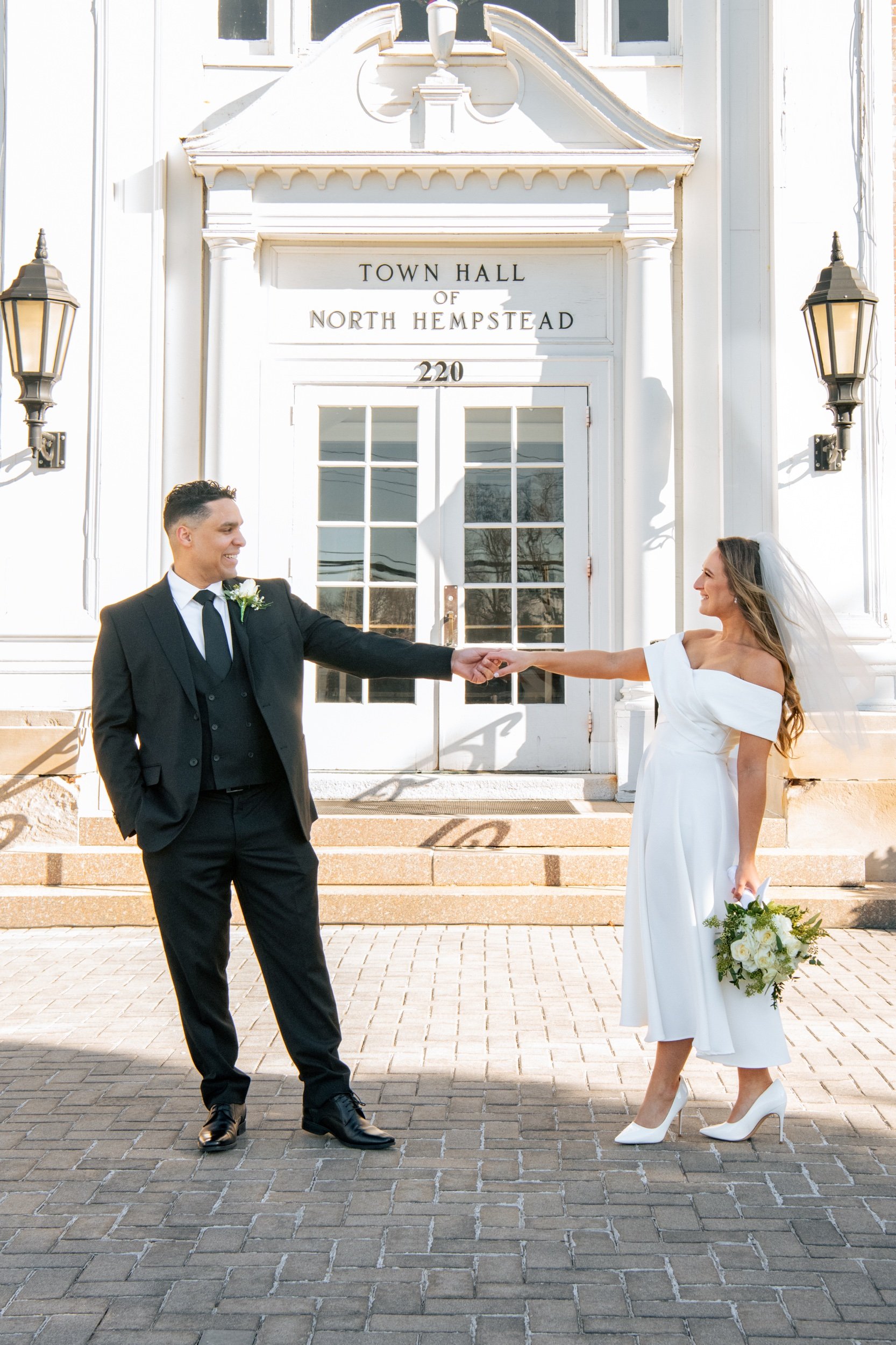 A groom in a black suit and a bride in a white dress holding a bouquet, holding hands outside a white town hall building with double glass doors, during a wedding ceremony.