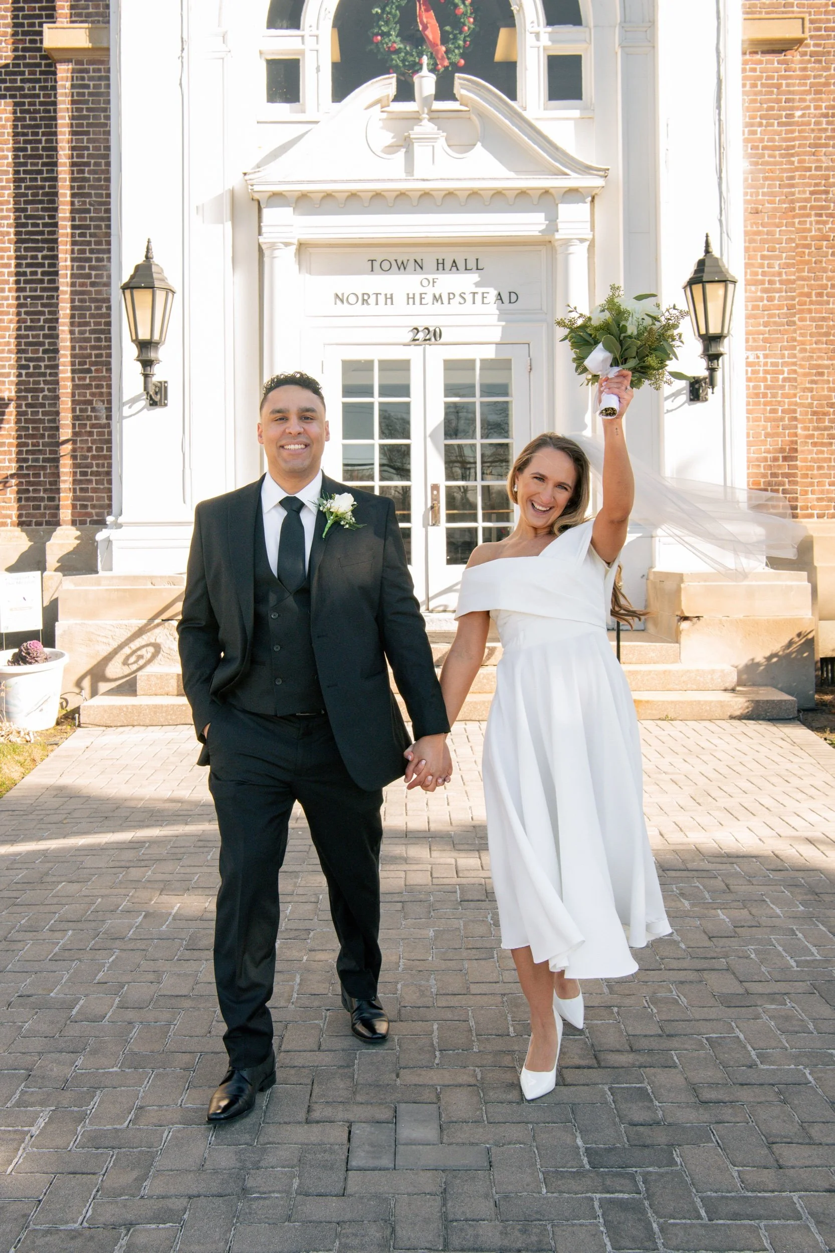 A joyful bride and groom holding hands and smiling outside the Town Hall of North Hempstead. The bride is wearing a white dress and holding a bouquet, while the groom is in a black suit and tie. The bride raises her bouquet in the air.