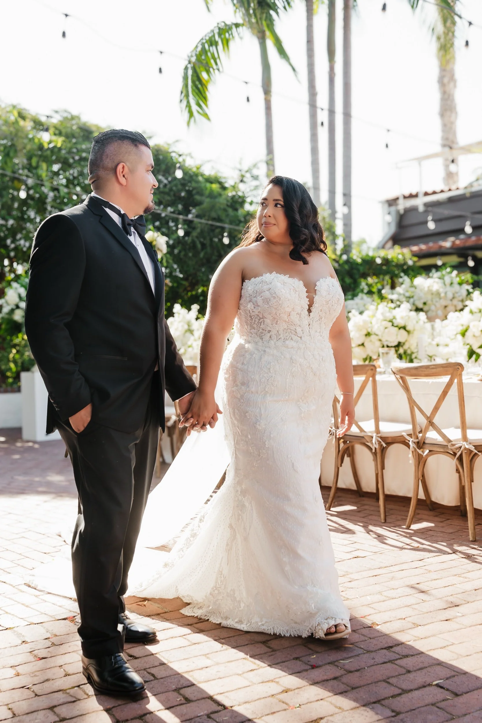 A bride and groom holding hands at their outdoor wedding ceremony, with white flowers and string lights in the background.