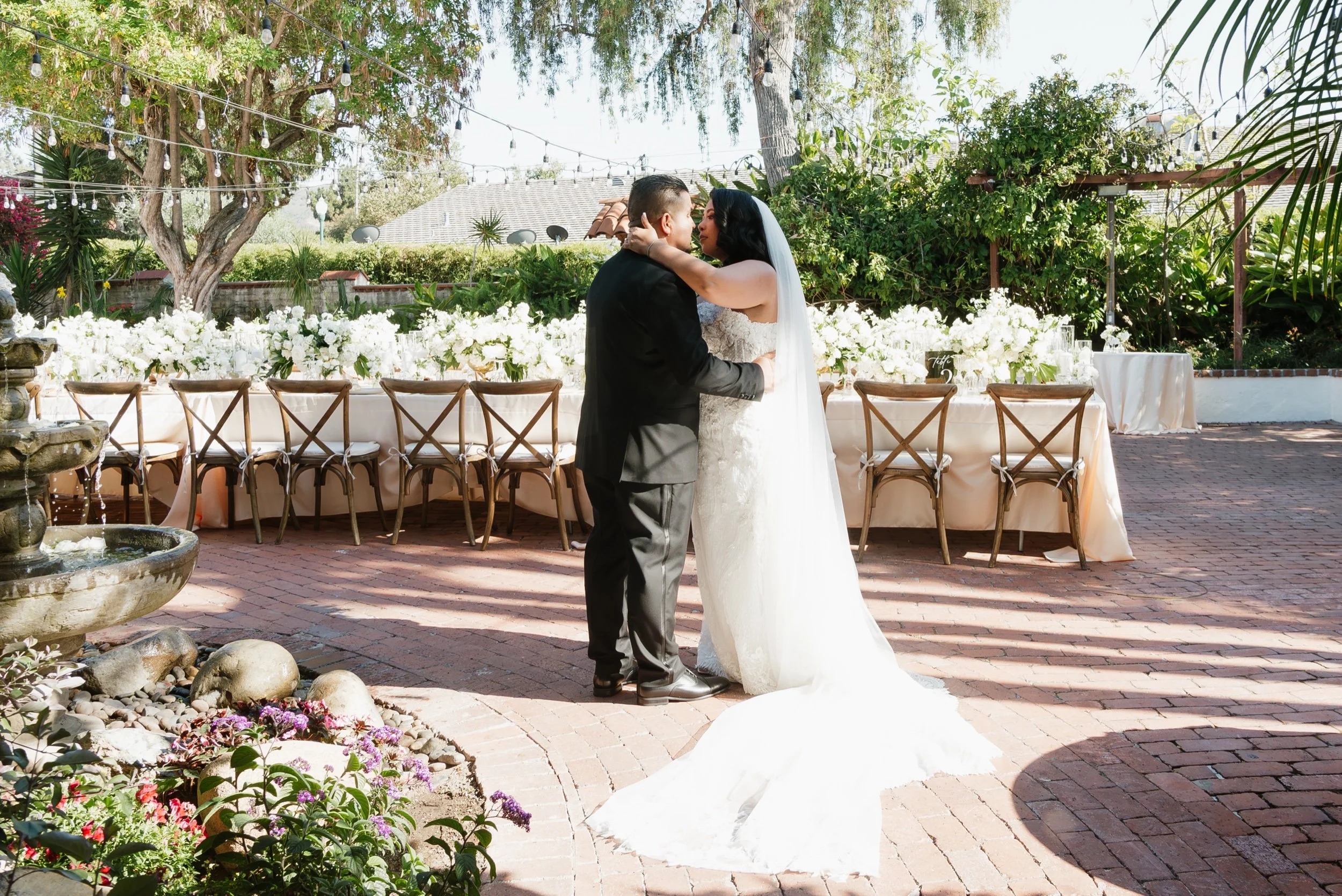 A bride and groom sharing a dance or embrace during their wedding reception outdoors. The bride is wearing a white wedding gown with a veil, and the groom is in a black suit. They are in front of a decorated table with white flowers, under trees with