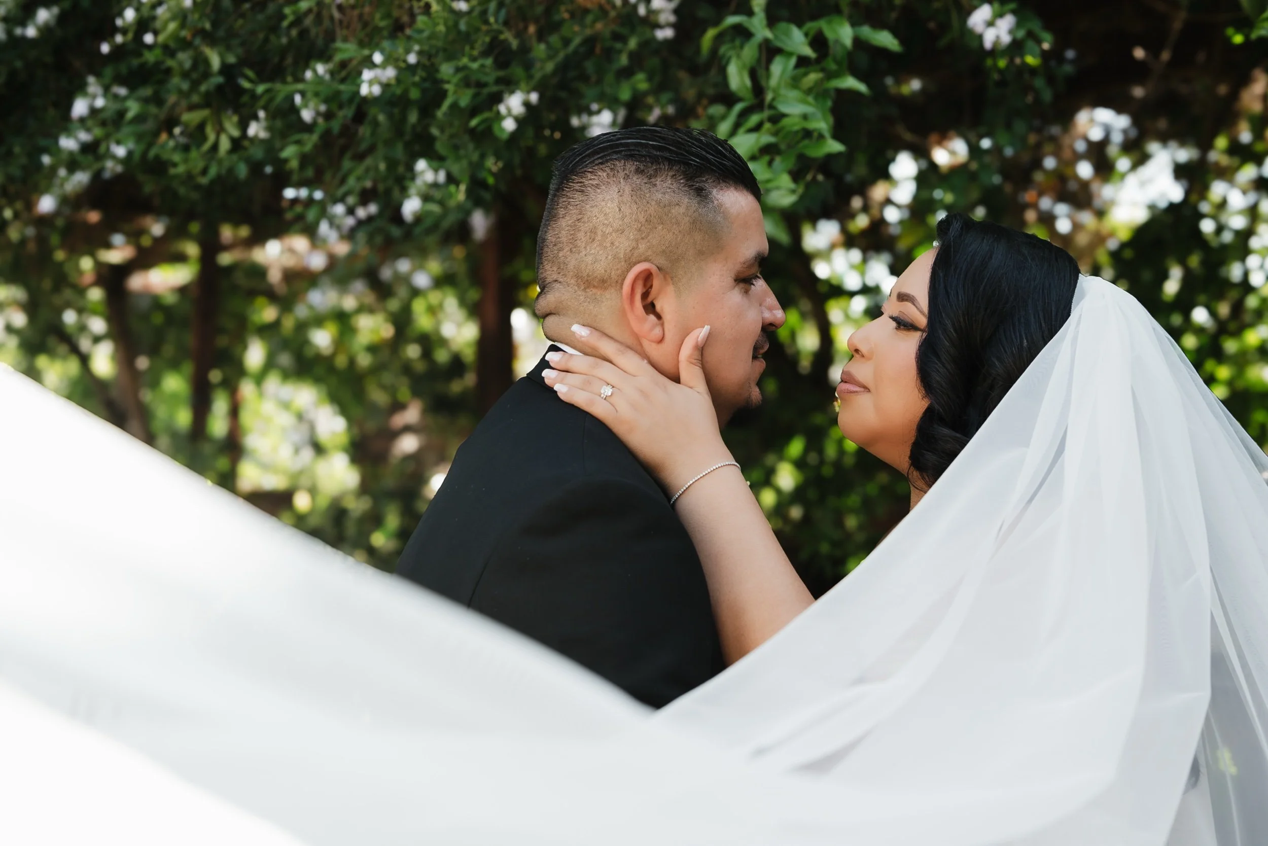 A couple dressed in wedding attire gazing into each other's eyes outdoors with greenery in the background.
