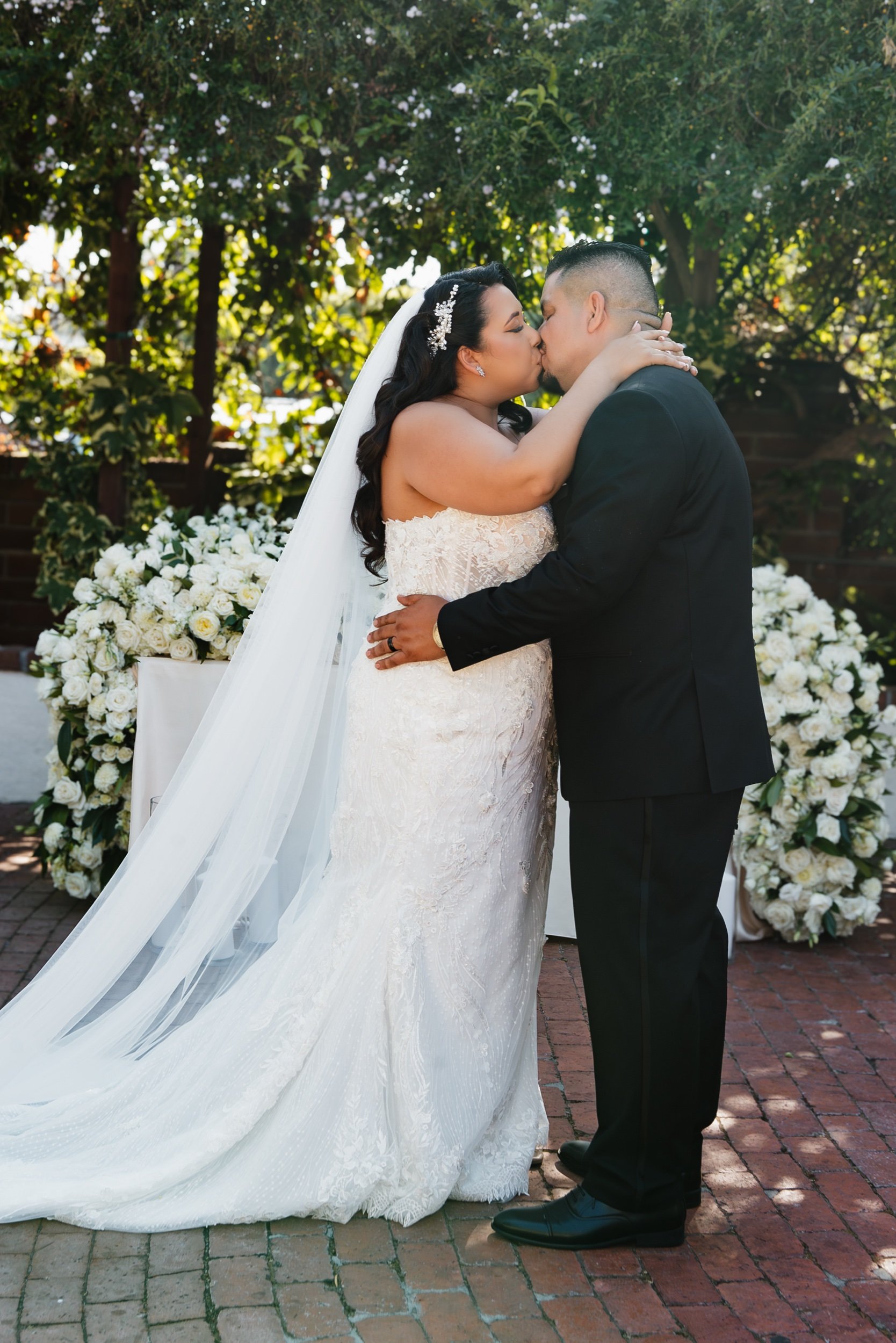 A bride and groom kiss during their wedding ceremony outdoors, with flowers and greenery in the background.