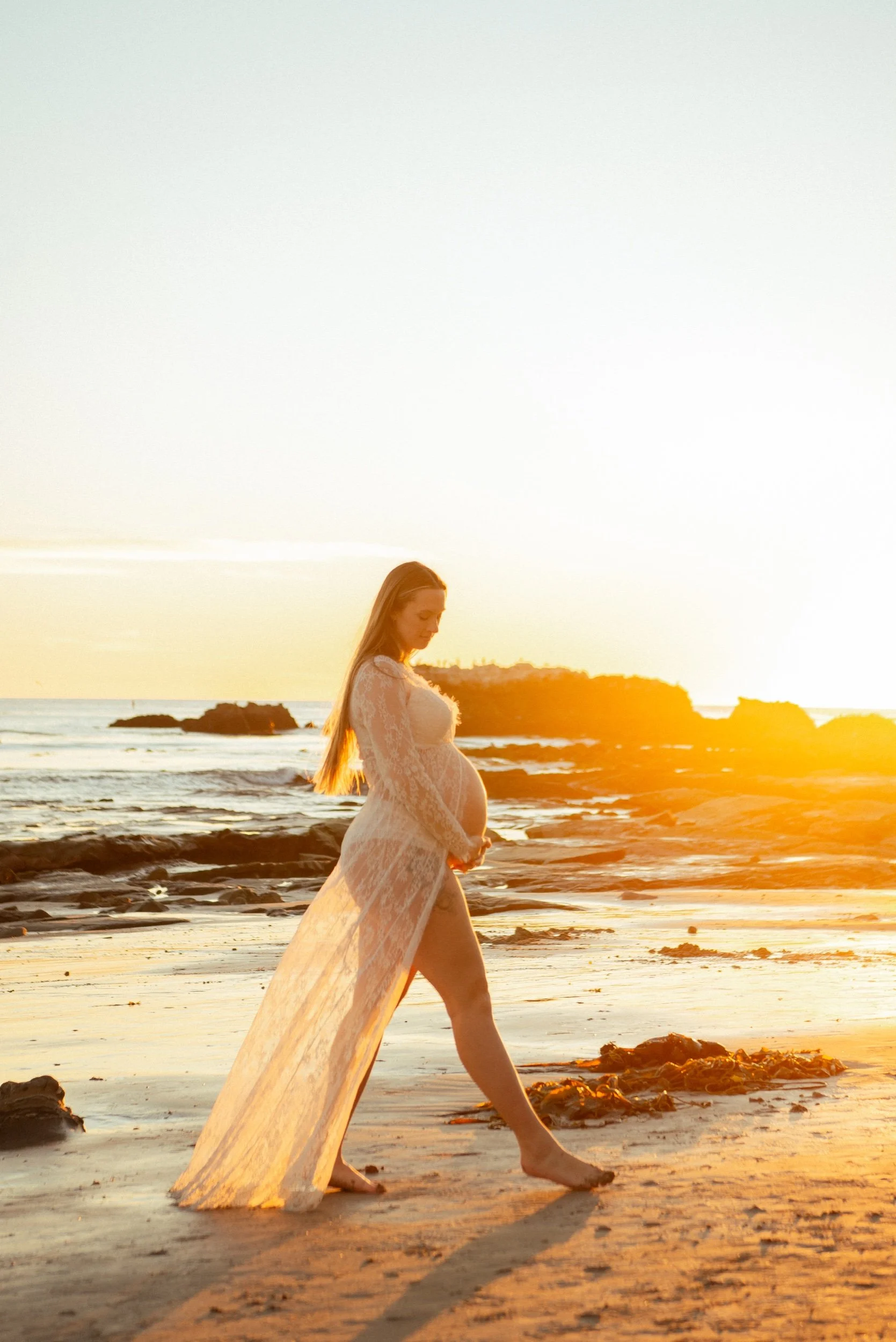 Pregnant woman in a long, white lace dress walking along the beach at sunset.