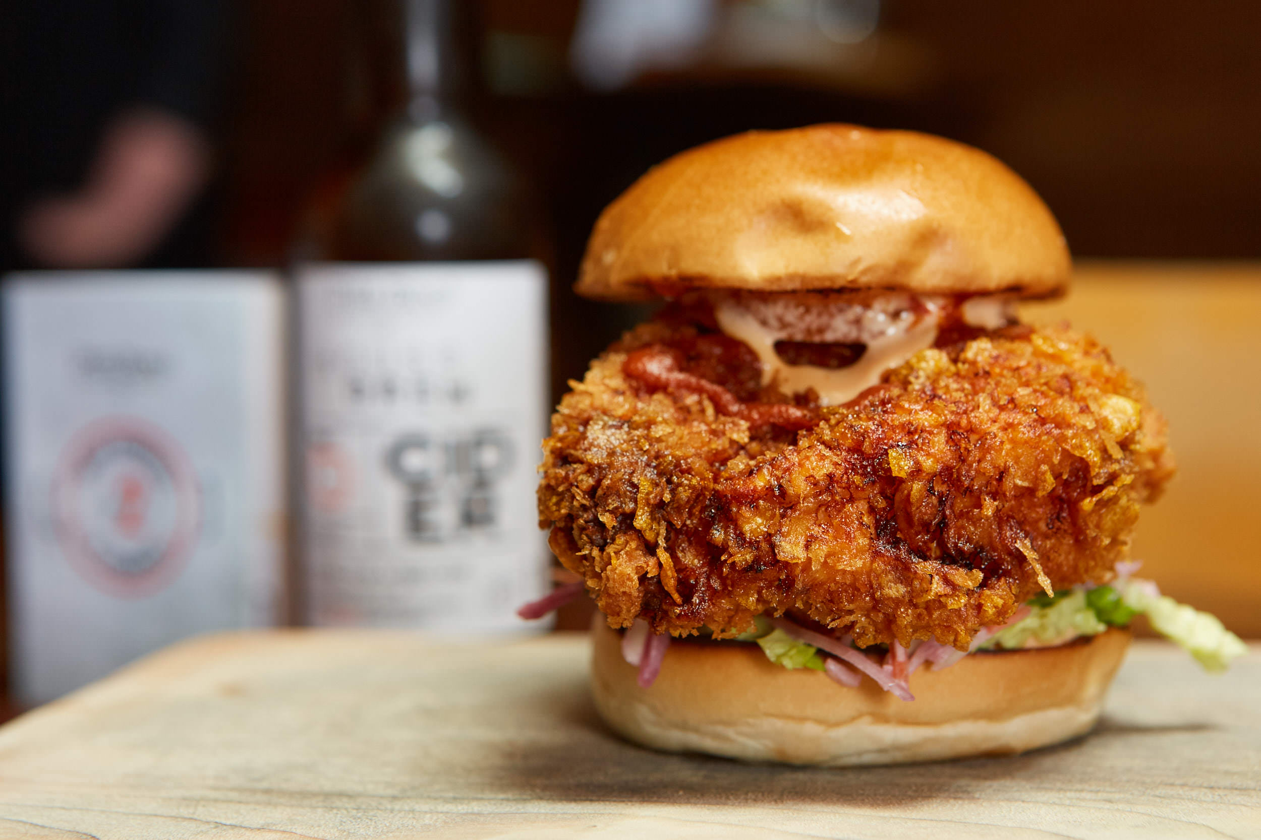 A crispy fried chicken sandwich with lettuce, tomato, and mayonnaise, topped with a soft bun, placed on a wooden surface. In the background, there are two bottles, one labeled 'O' and the other 'G'.