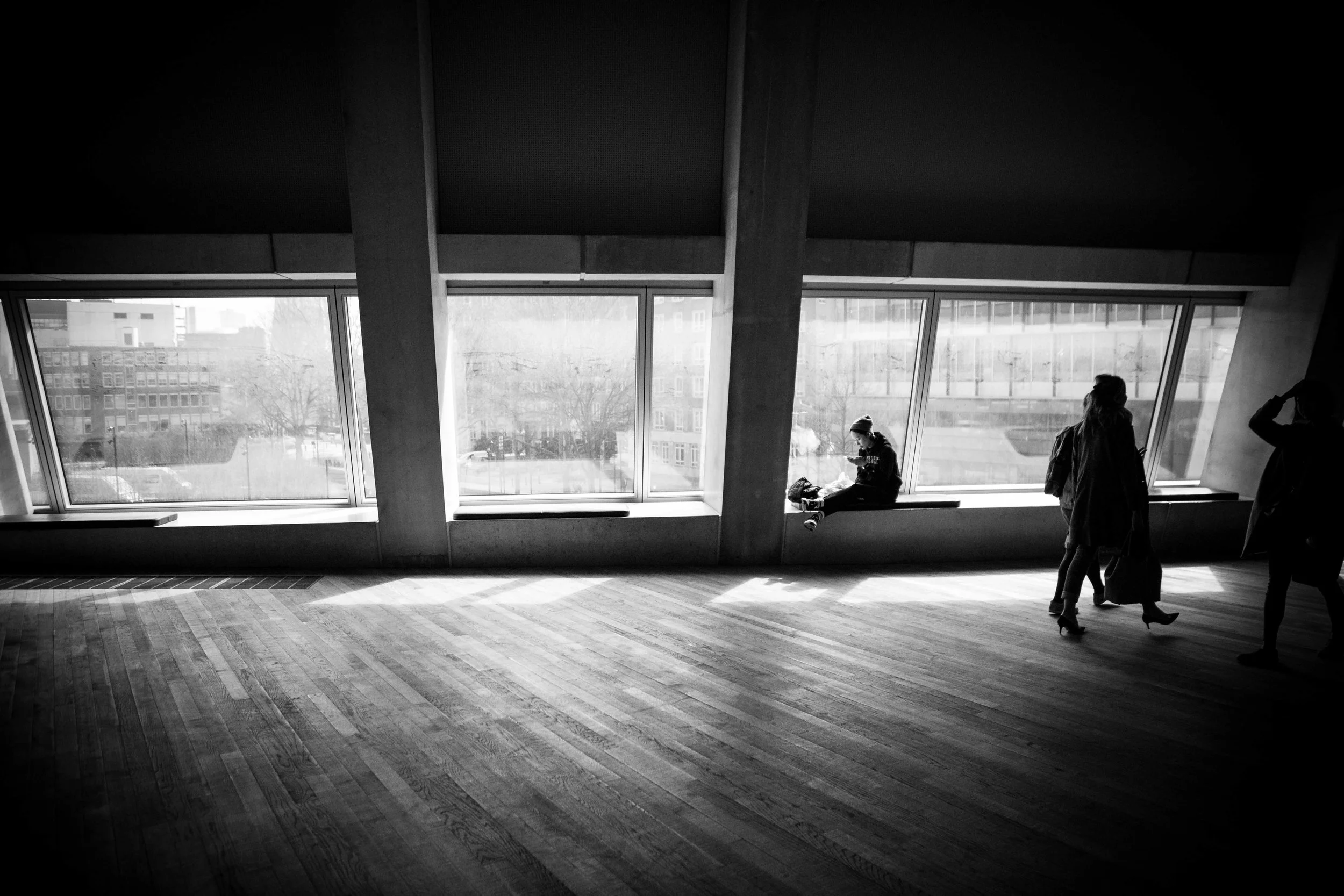 Silhouette of three people standing inside a building with large angled windows, overlooking city buildings and trees outside. One person is sitting on a window ledge, reading or looking at something, while two others are standing, one with a hand ra