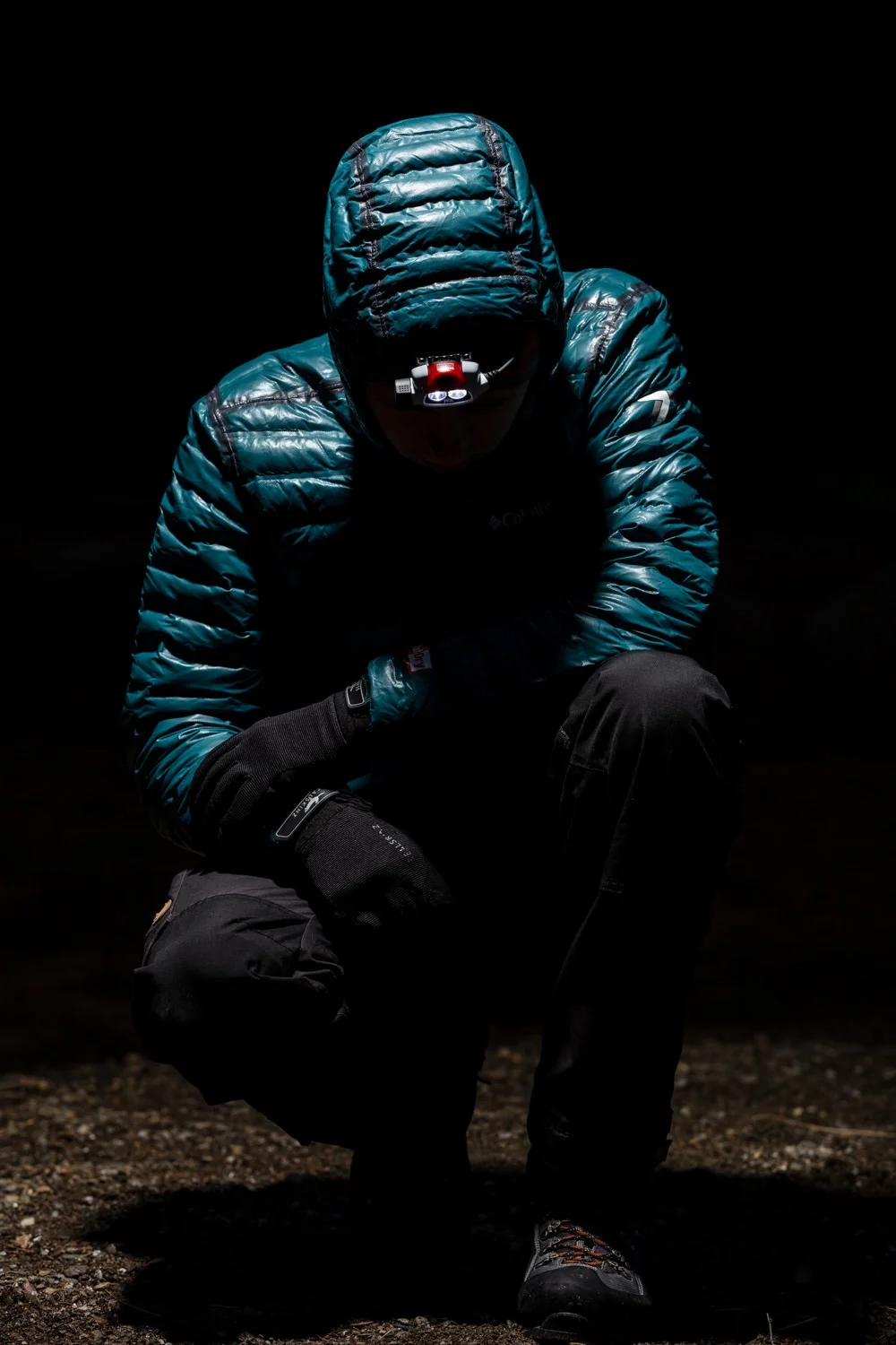 Person in outdoor gear, wearing a puffy jacket with his hood up, crouching in darkness with headlamp turned off.