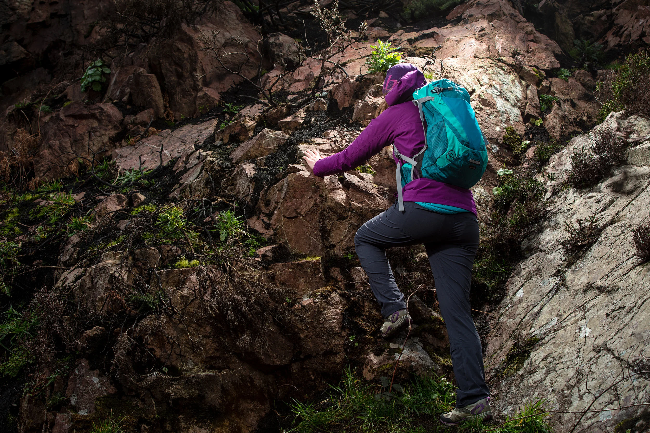 A person wearing a purple jacket, black pants, and gray hiking boots with purple accents is climbing a rocky outdoor slope, carrying a turquoise backpack and wearing a purple hat.
