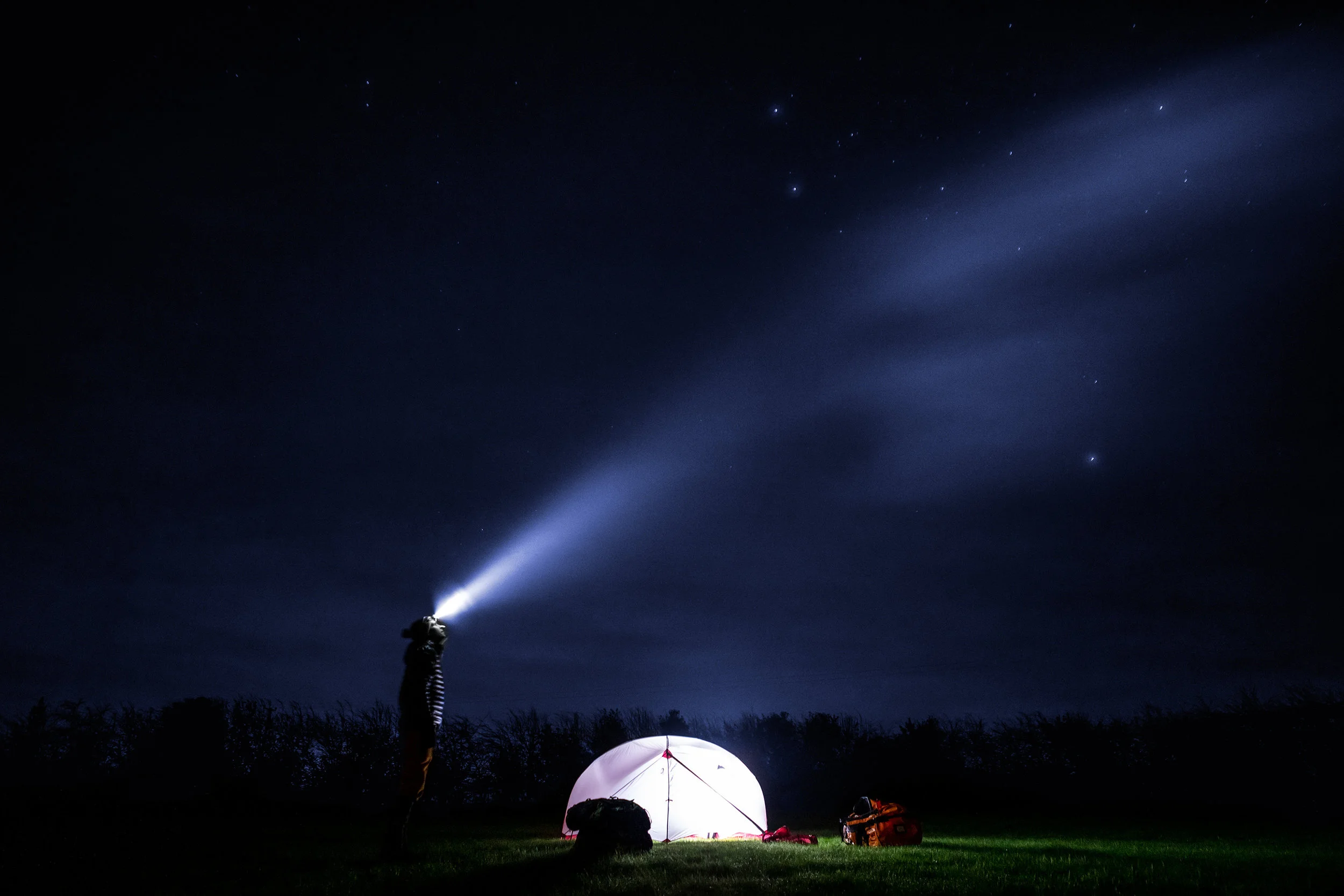 Person camping on grass at night with a starry sky, a star projector or flashlight, and a backpack.