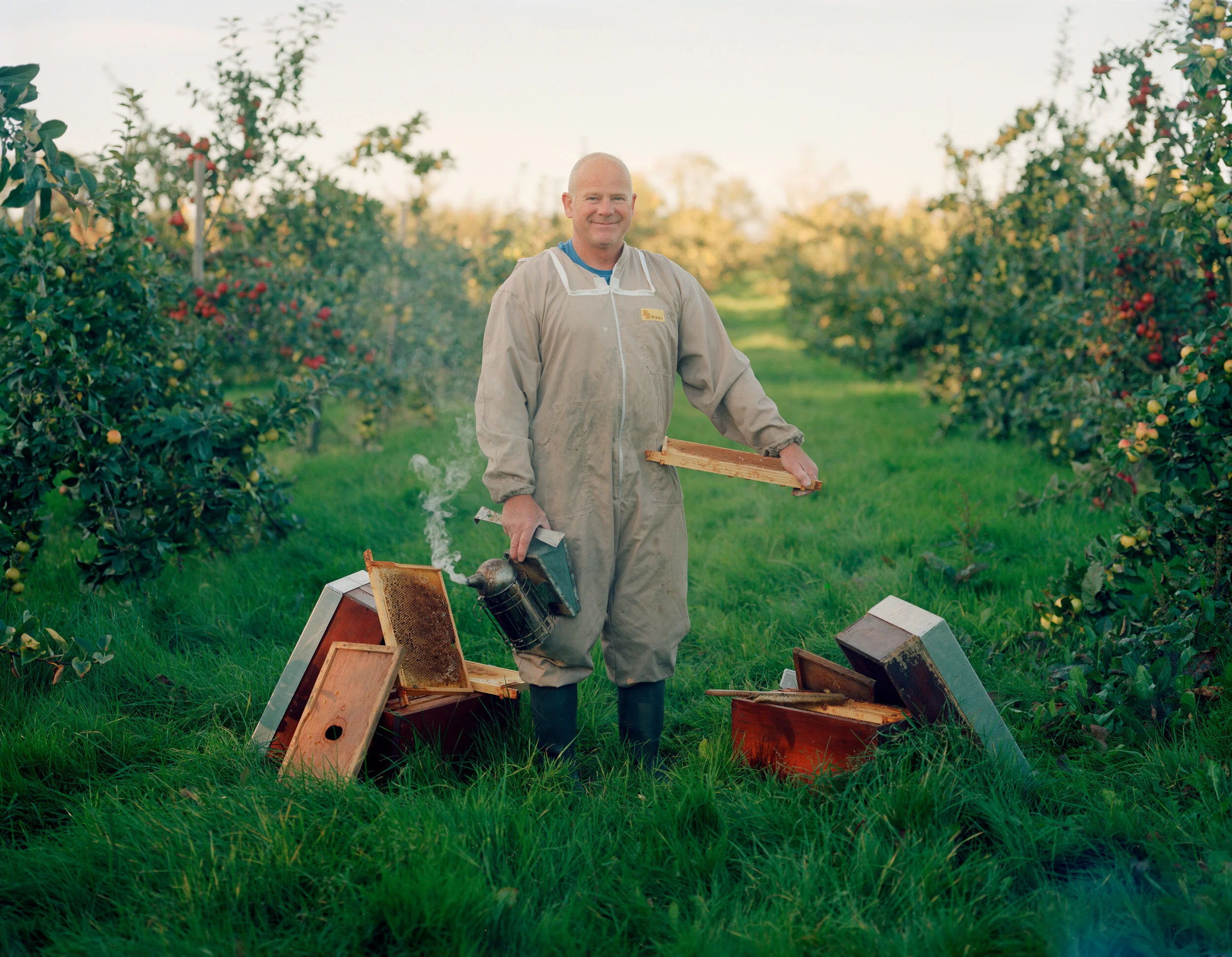 A man smiling at the camera in an apiary with honeybees and hive boxes around him, holding a frame from a beehive.