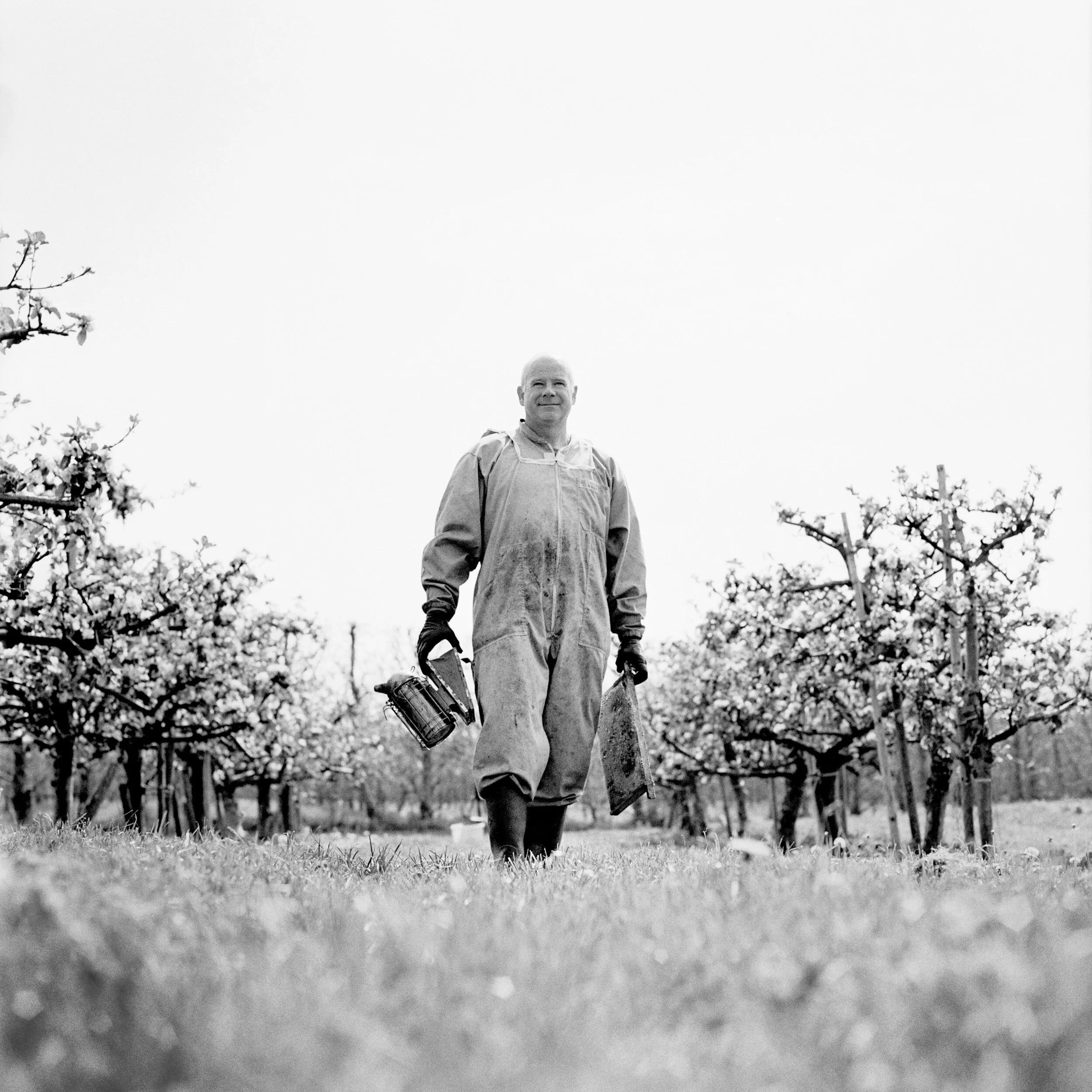 A man with a bald head wearing a jacket and gloves walking outdoors in a black-and-white photo, looking ahead, with trees and a fence in the background.