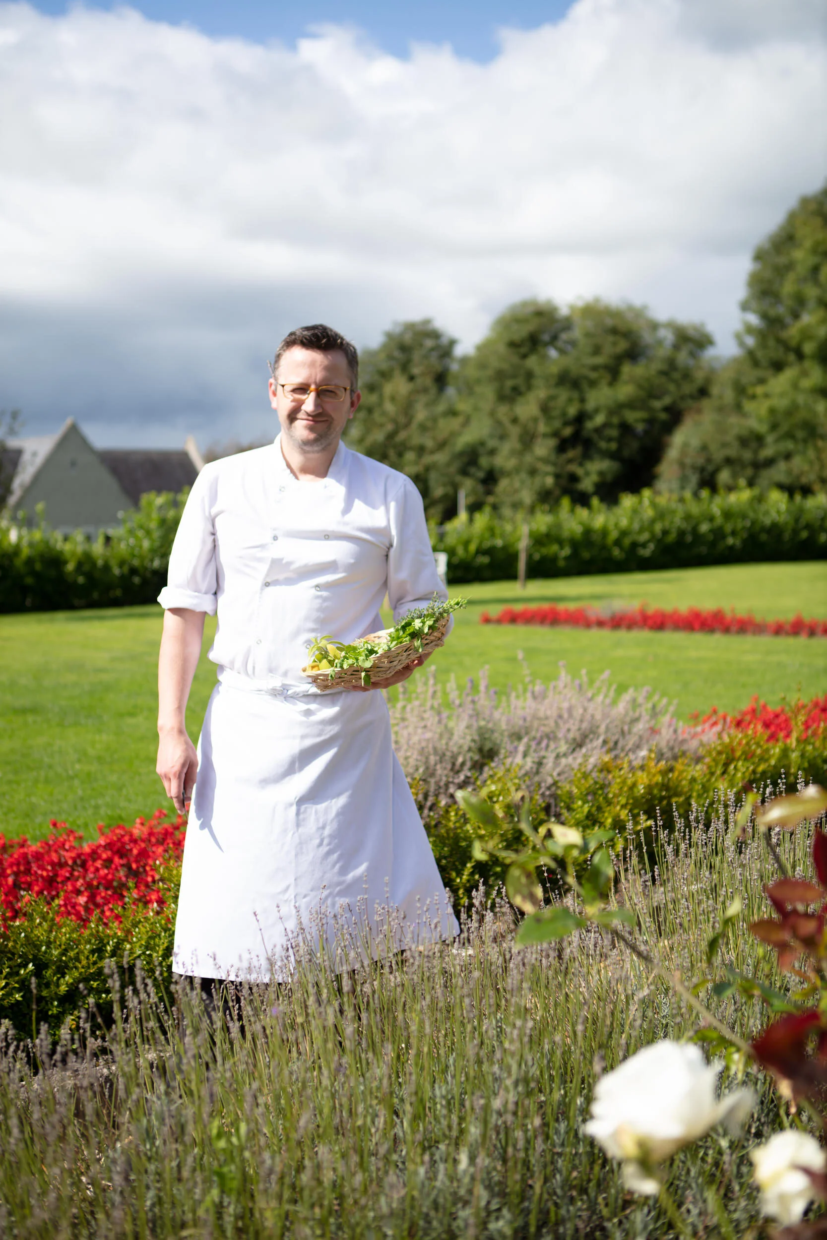 A man dressed as a chef standing in a garden with colorful flowers, holding a basket of greens, with a cloudy sky and trees in the background.