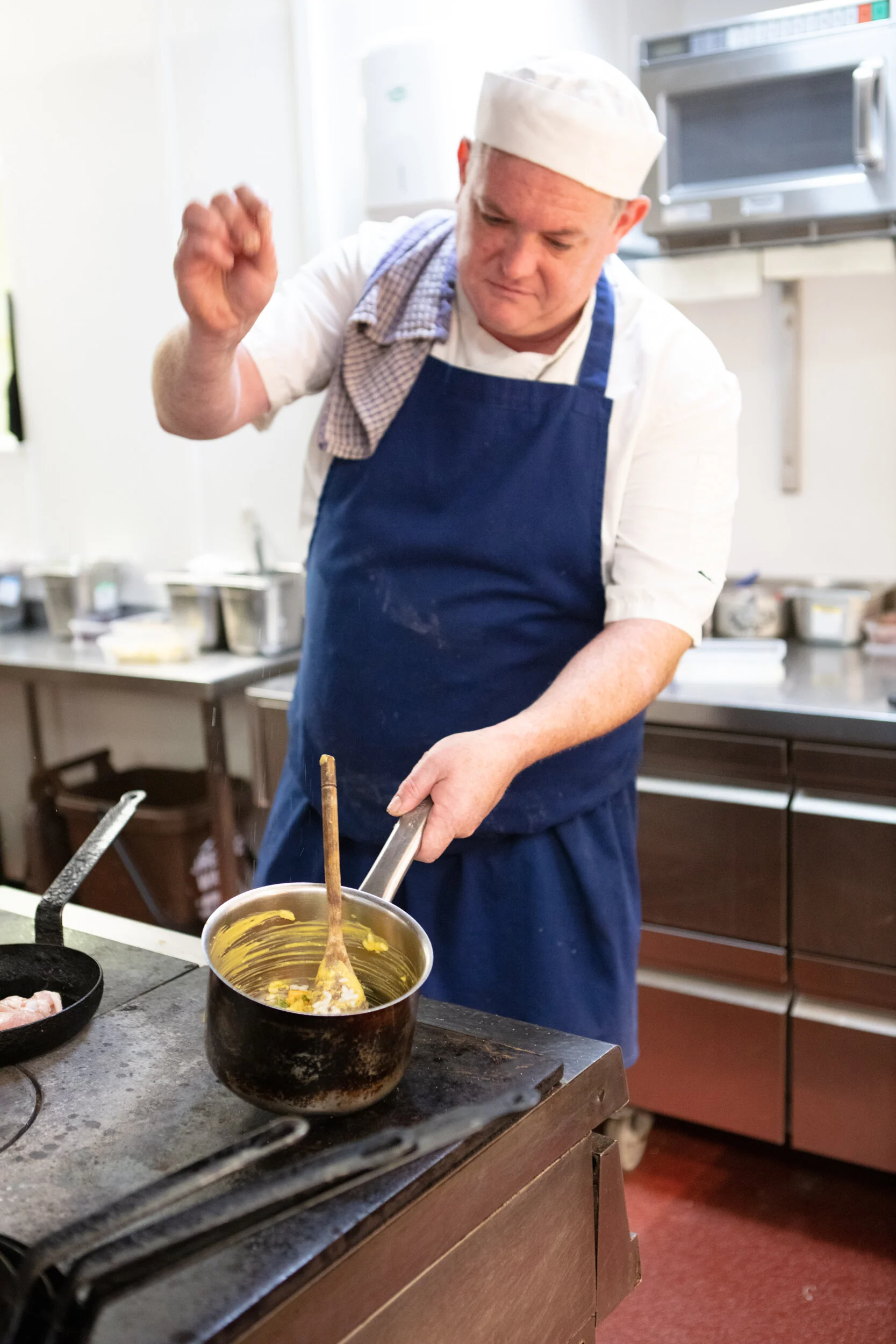 A chef wearing a white hat, white shirt, and blue apron is cooking on a stove, stirring a pot with a wooden spoon in a kitchen.