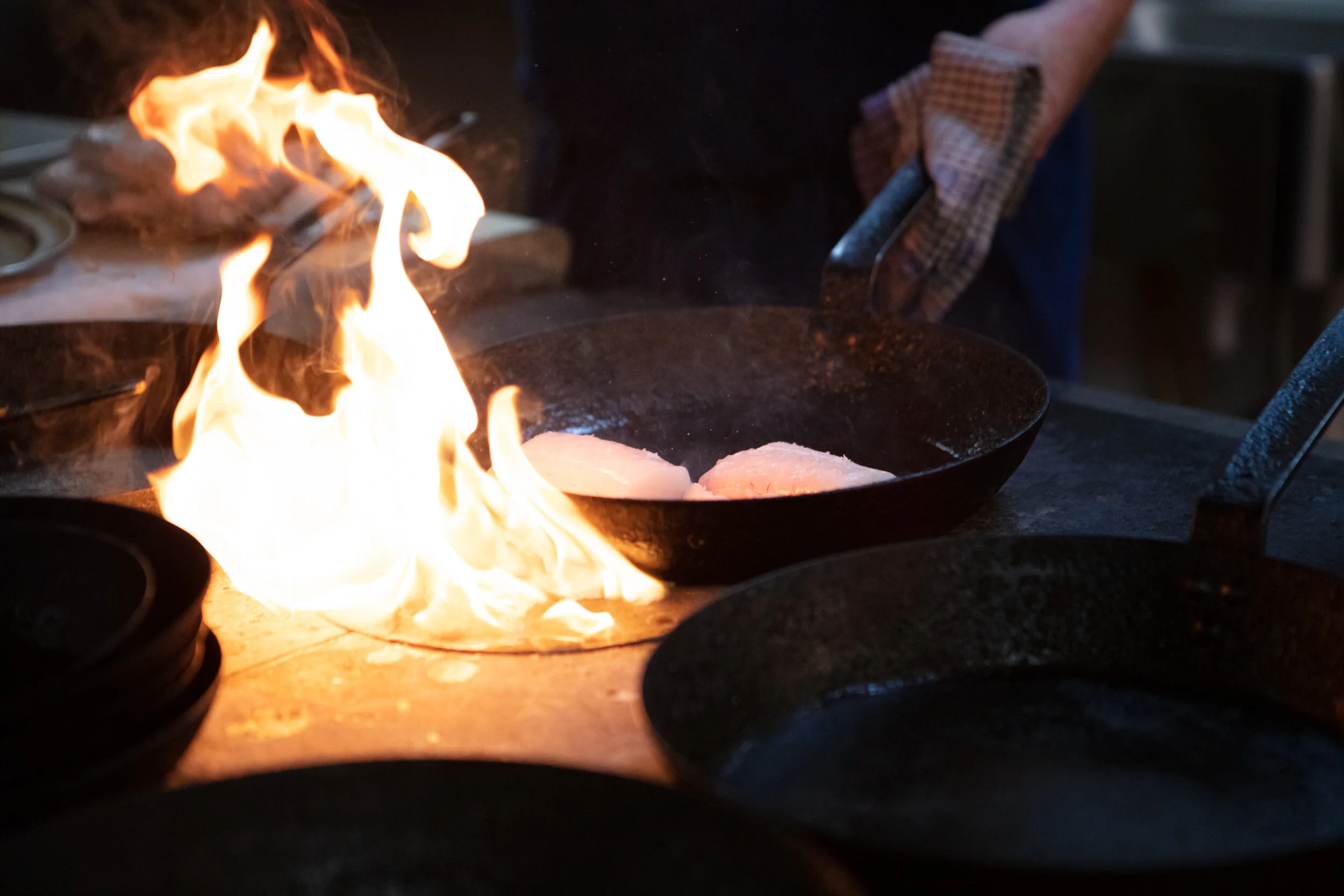 The image shows a close-up of a kitchen scene with flames coming out of a pan on the stove. A person wearing an oven mitt is holding a spatula or utensil near the stove, with a dark background indicating a kitchen environment.