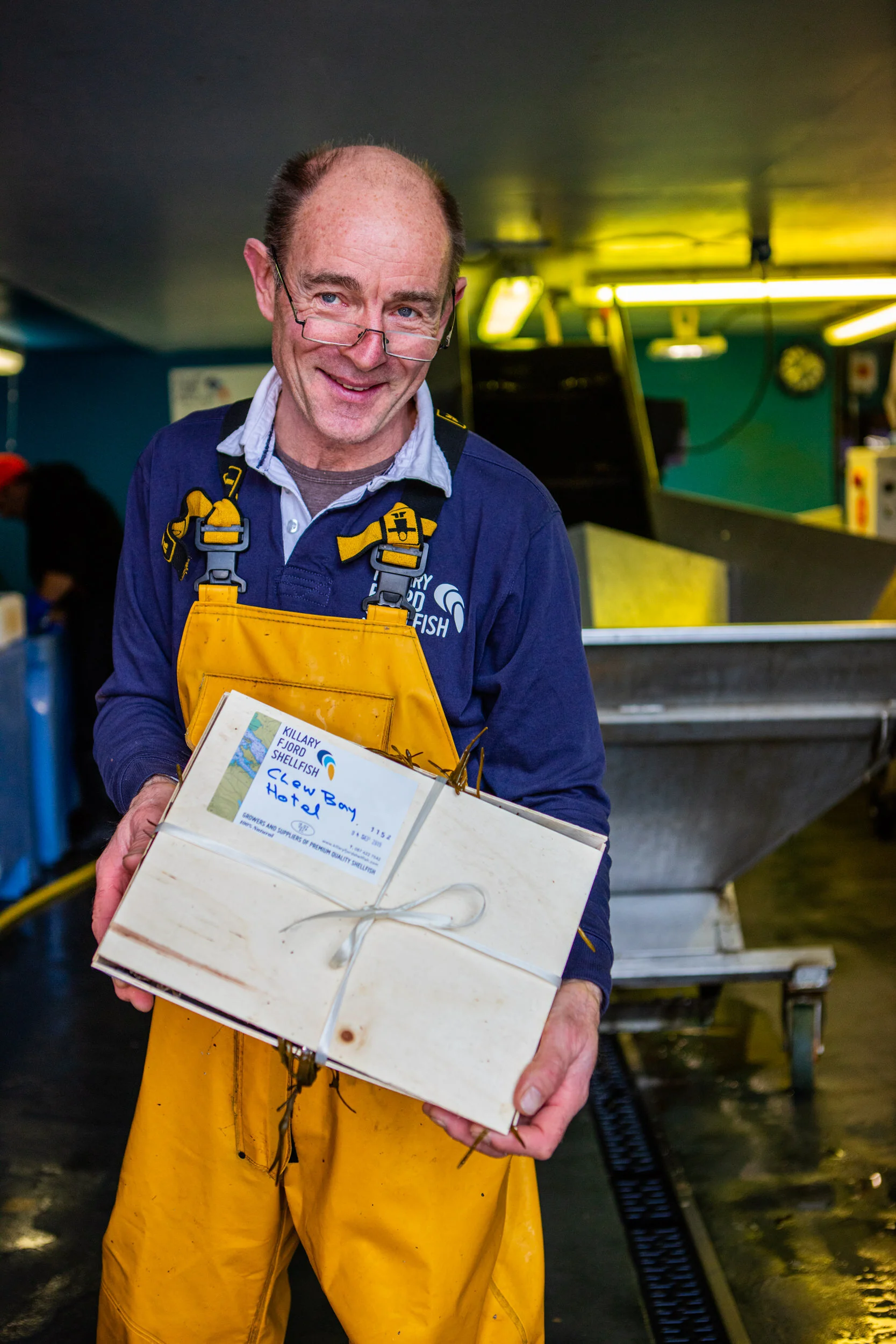 An older man wearing yellow waterproof overalls and glasses, smiling, holding a white box tied with a string. The box has a label indicating it is from Killary Fjord Shellfish and is addressed to Clwyd Bay Hotel.